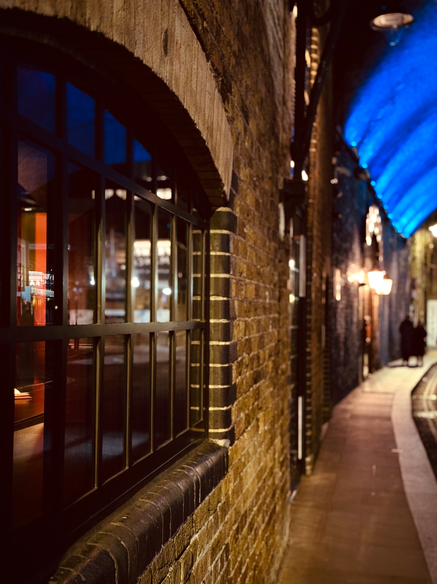 Nighttime scene of a brick building with a barred window and a blue awning, with two people walking on the sidewalk in the distance.