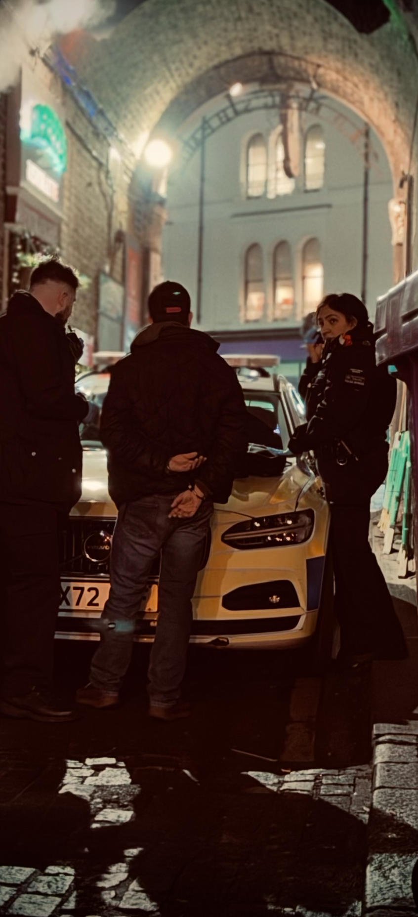 Three people, including a female officer, are standing around a yellow car in a dimly lit urban alley. The officer appears to be talking to one of the men, while another man looks at his phone. The background features brick walls and arched windows.