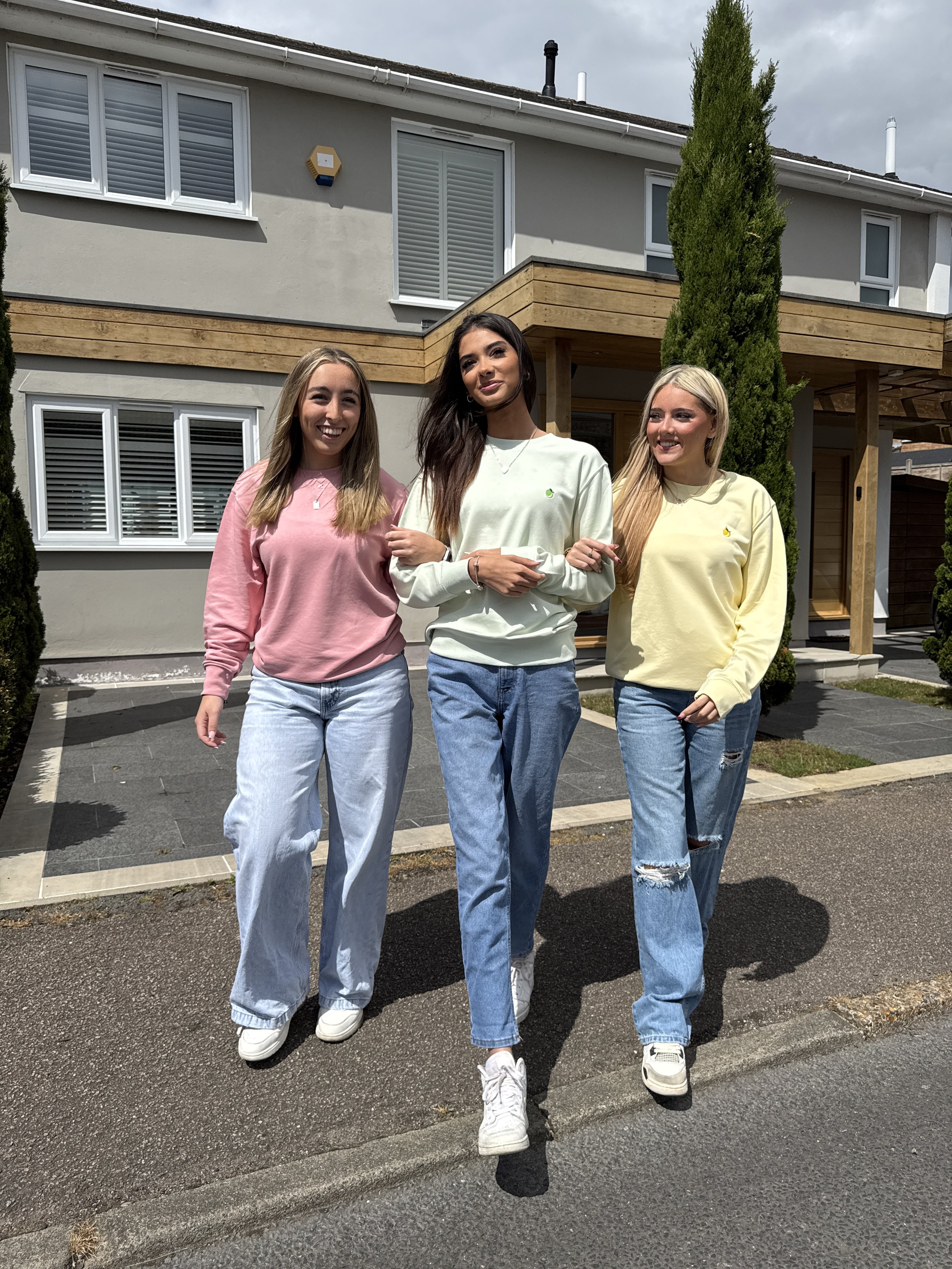 Three young women standing together outdoors in front of a modern house with large windows and trees, smiling and posing for a photo.