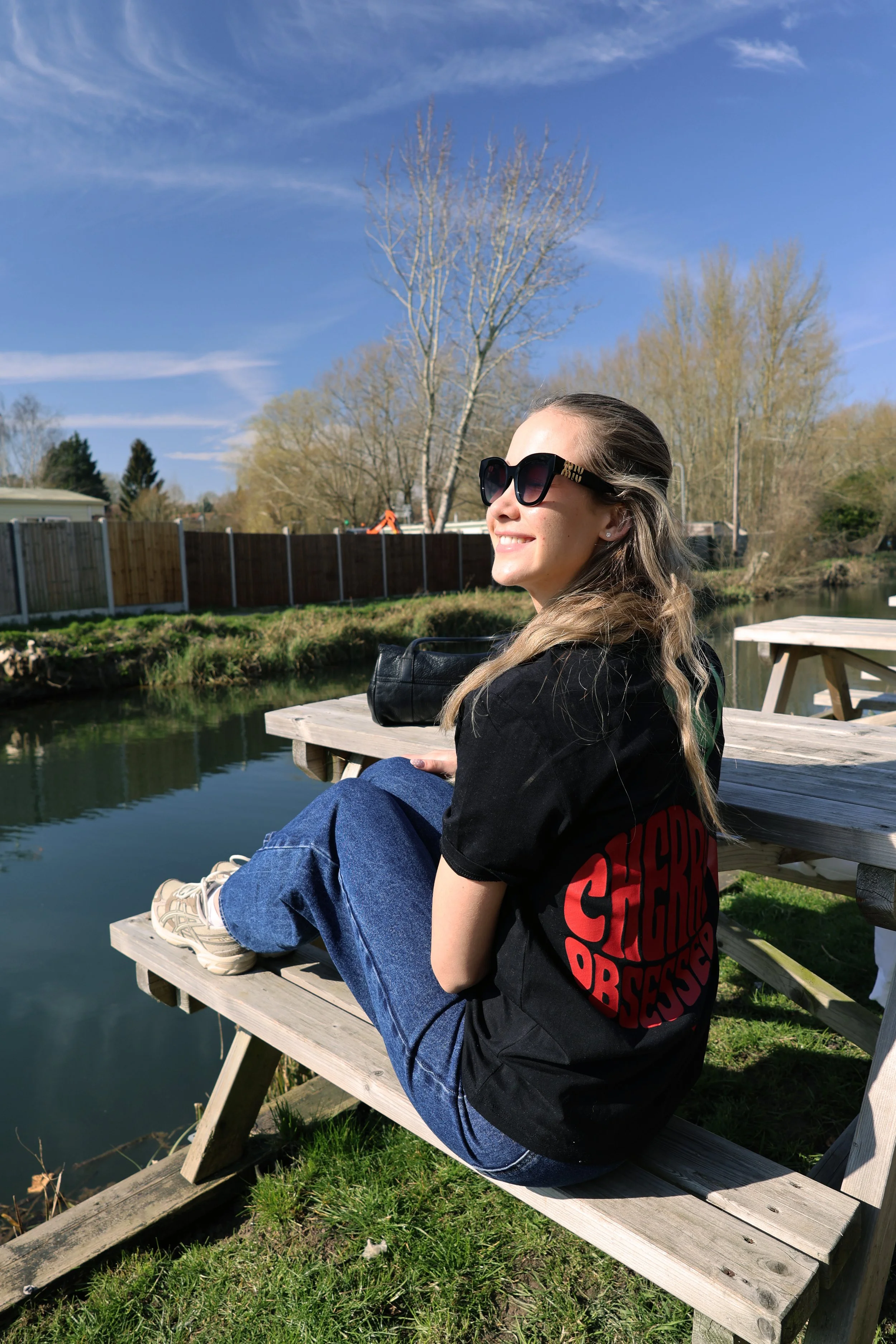 A young woman with long blonde hair wearing dark sunglasses and a black t-shirt with red print, sitting on a wooden bench by a small pond, smiling and enjoying a sunny day.
