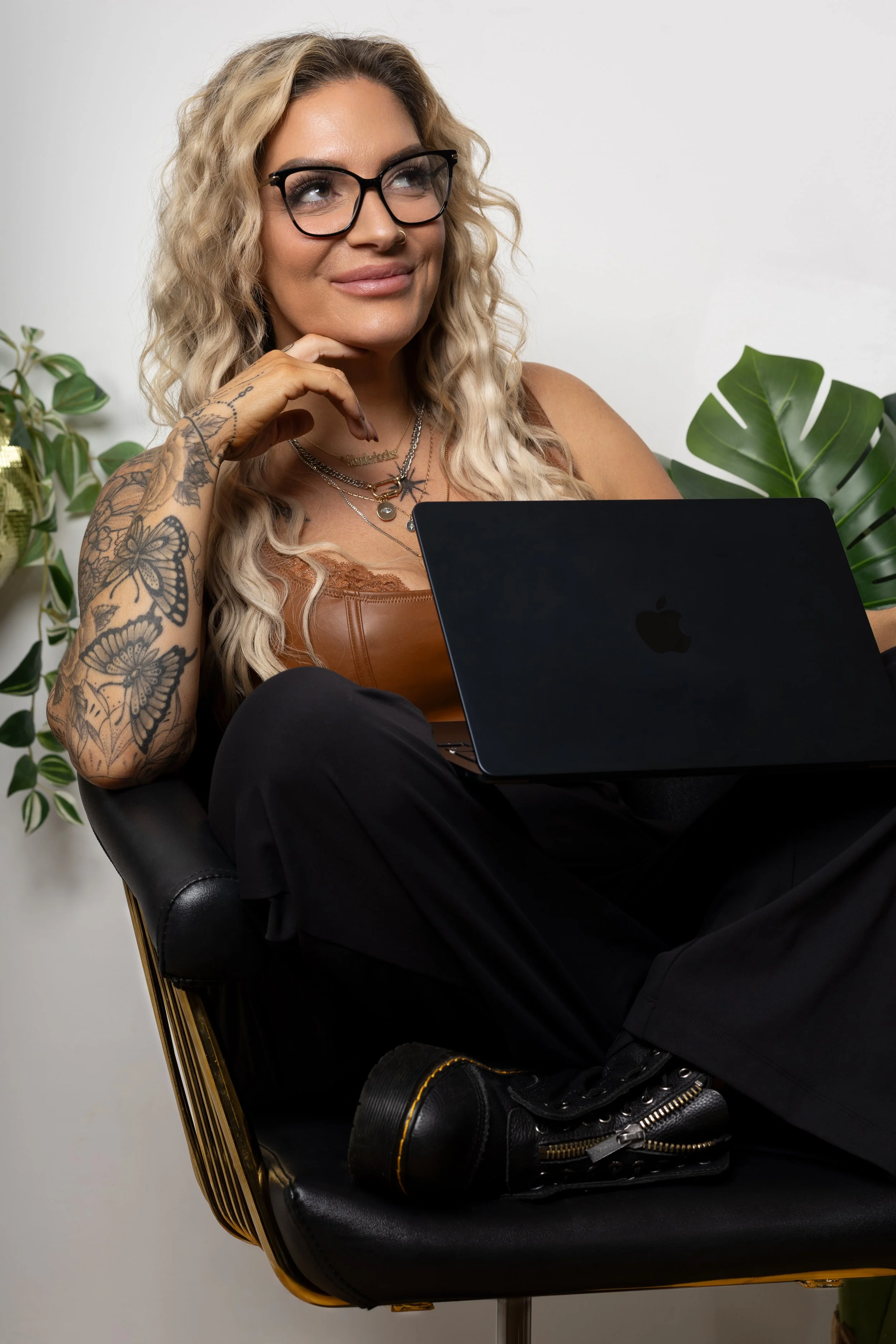 A woman with blonde curly hair, glasses, and tattoos on her arm is sitting on a black chair with gold accents, holding a black laptop on her lap. She is wearing a brown sleeveless top, multiple necklaces, and dark pants. There are green plants in the