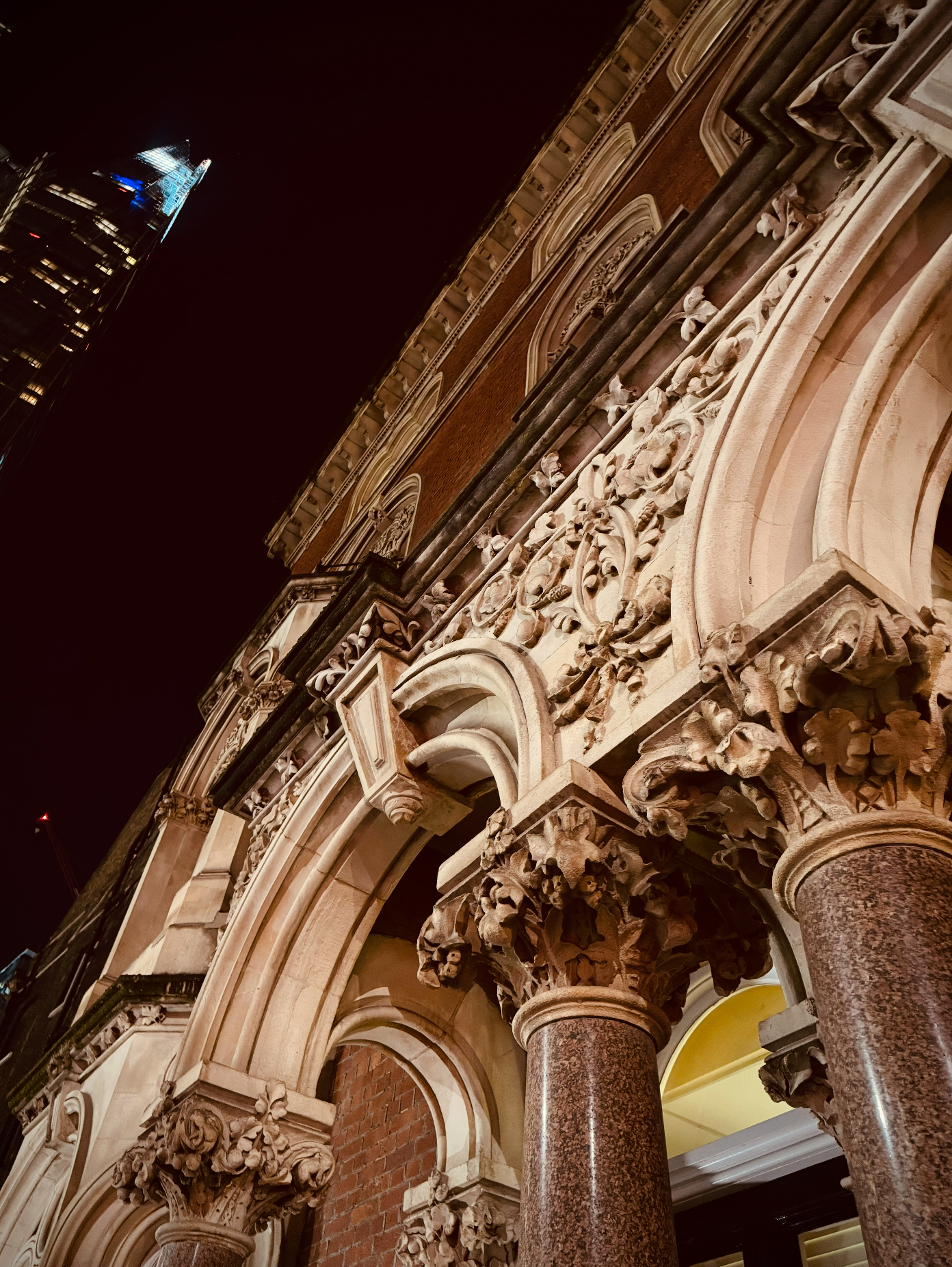 Close-up of ornate architectural details on a building facade, featuring carved stone columns and decorative arches, with a modern skyscraper visible in the background at night.
