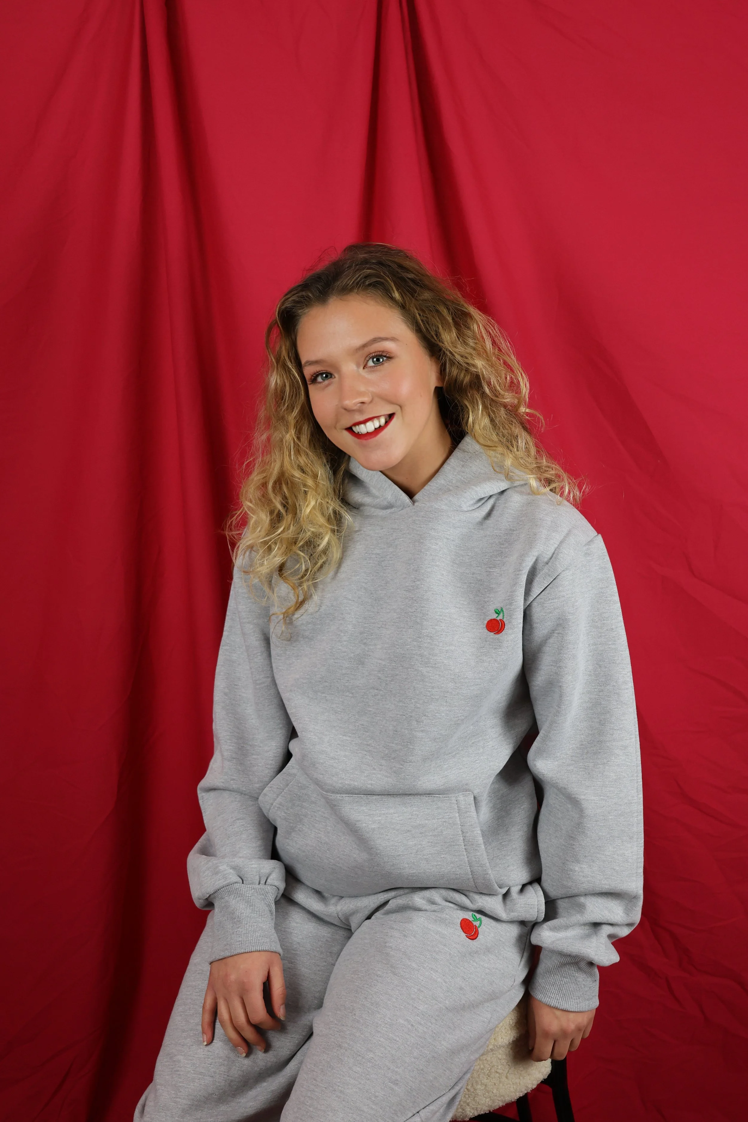 Young woman with curly blonde hair smiling, wearing a gray hoodie and sweatpants with small cherry embroidered logos, sitting against red draped fabric background.