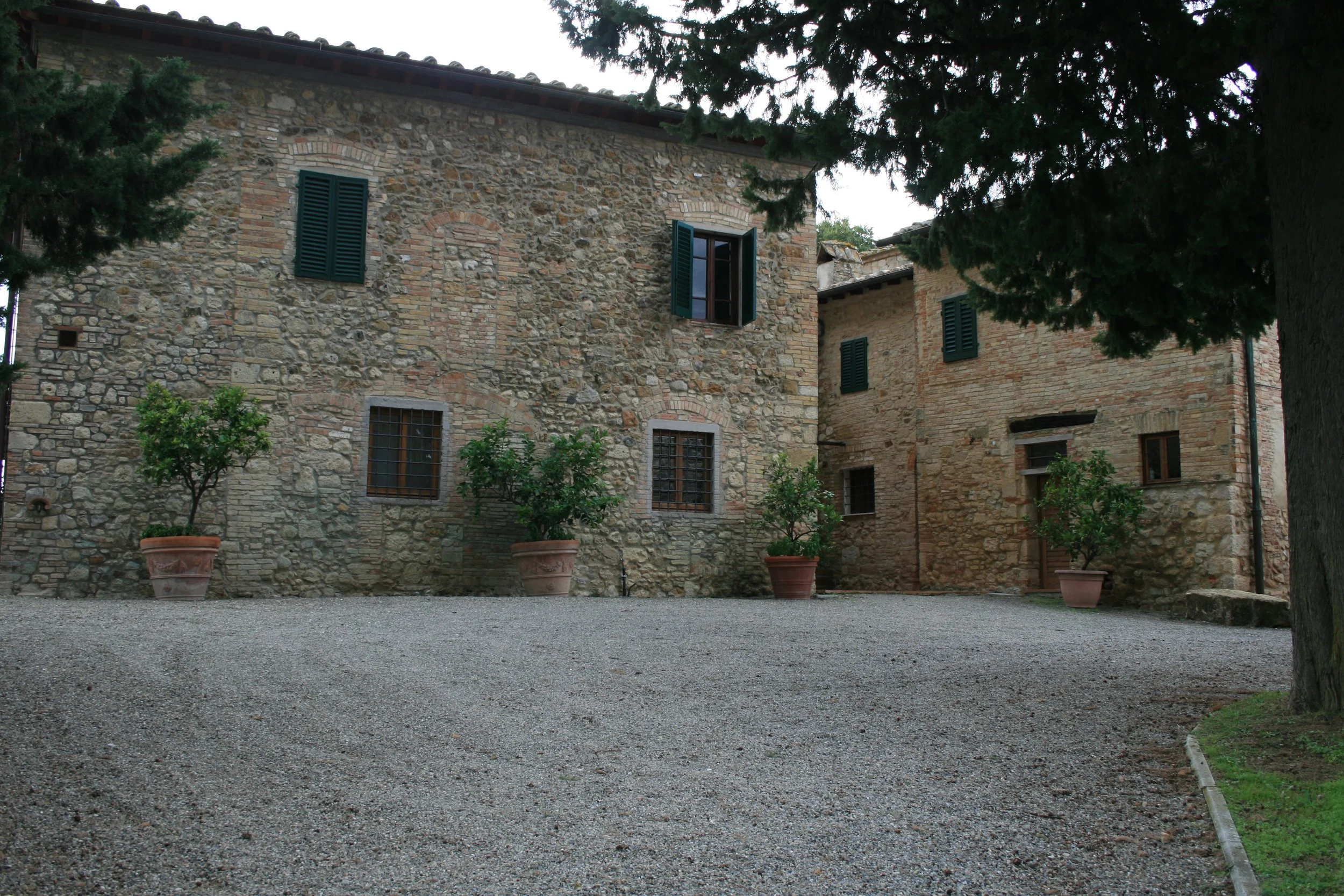 Historic stone building with green shutters and potted trees in front, surrounded by a gravel courtyard and large trees.