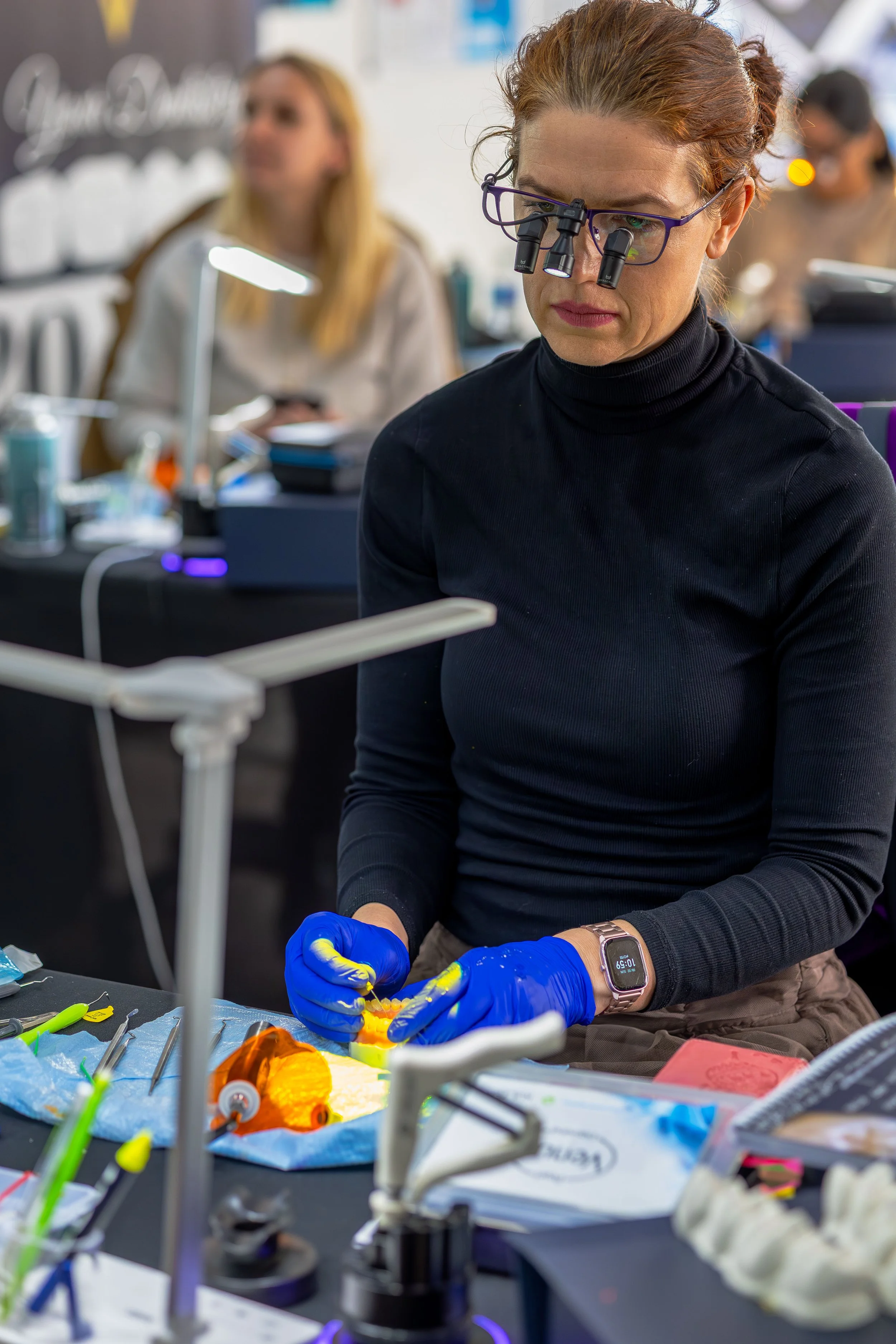 A woman wearing magnifying glasses with lights is examining a small object with blue gloves at a workspace with various tools and materials, with other people working in the background.