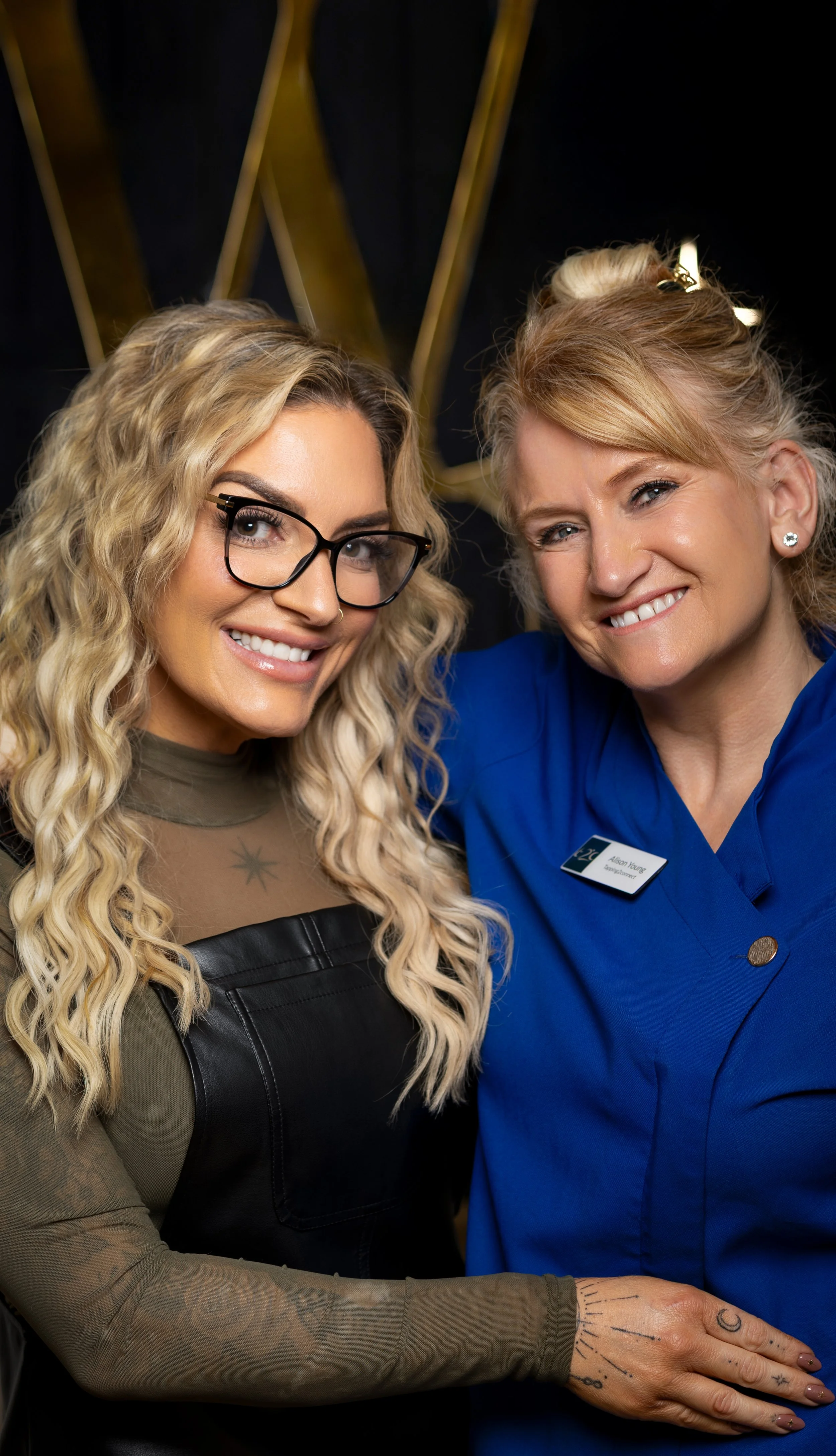Two women, one with glasses and tattooed hand, smiling and embracing each other, with a dark background and gold accents.