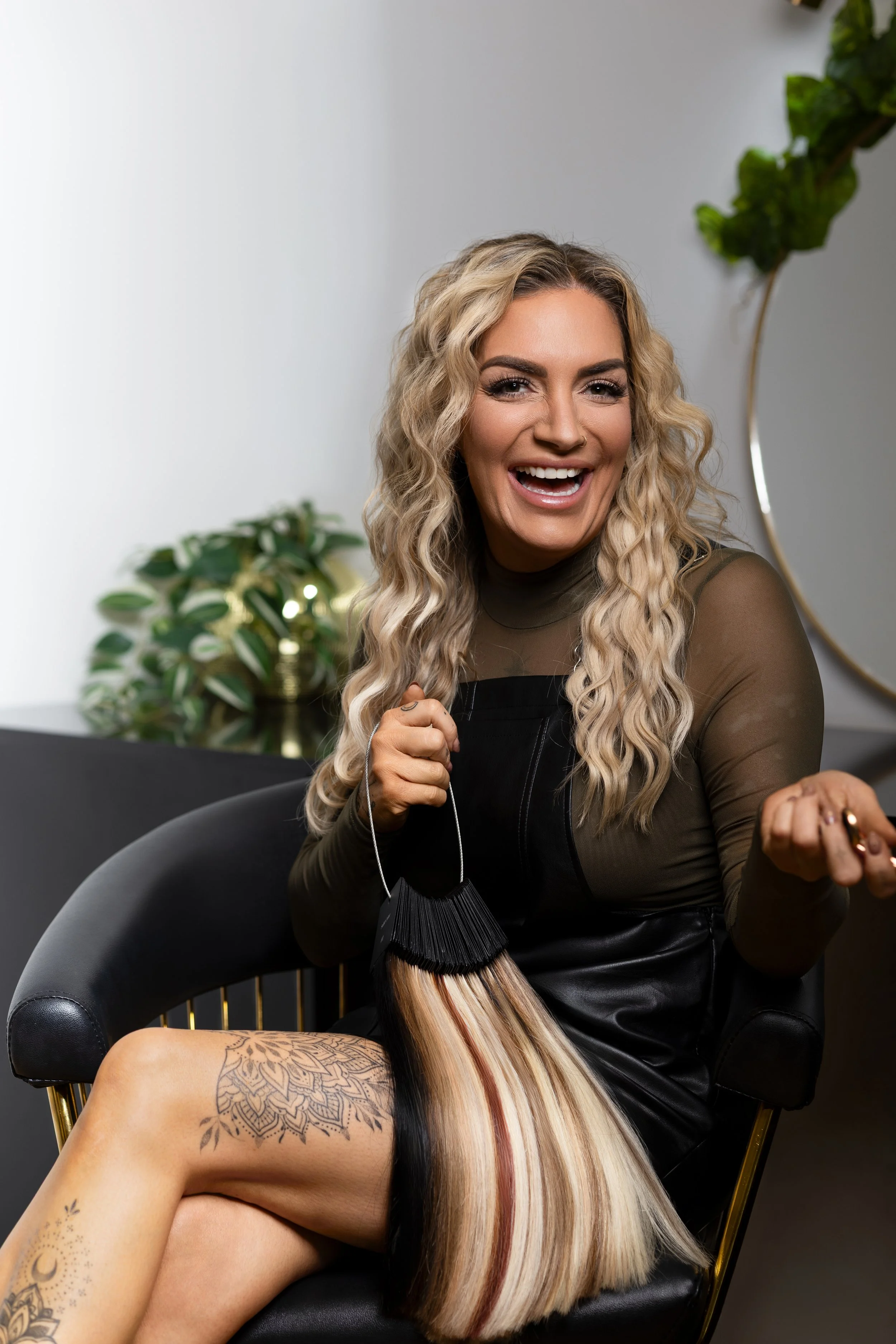 A woman with wavy blonde hair, tattooed legs, and a nose ring, sitting on a black chair and smiling while holding a hair color swatch.