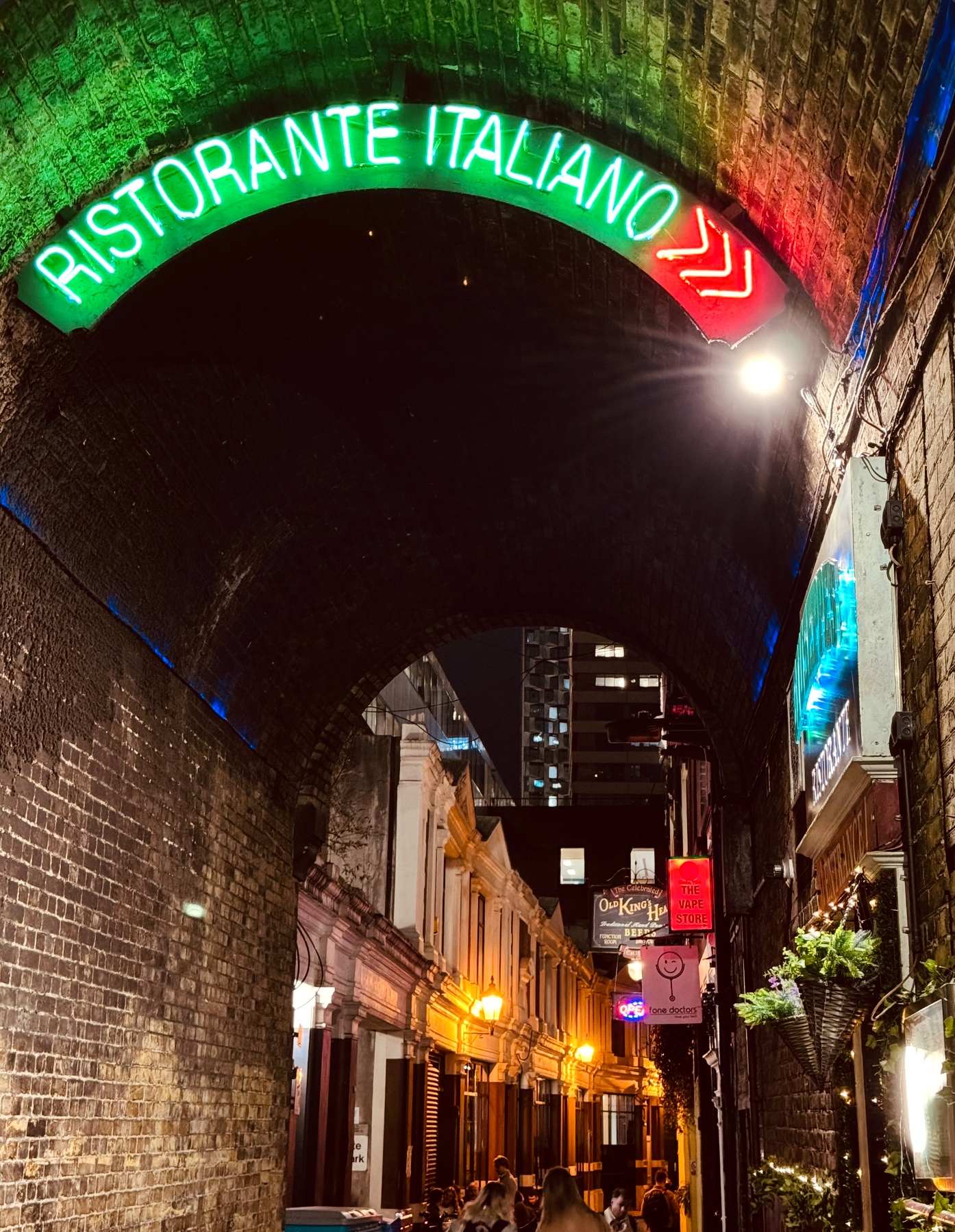 Night scene of a narrow street with brick buildings, illuminated by warm streetlights, under an archway with neon signs reading 'Ristorante Italiano' in green and red lights. People are walking and socializing along the street.