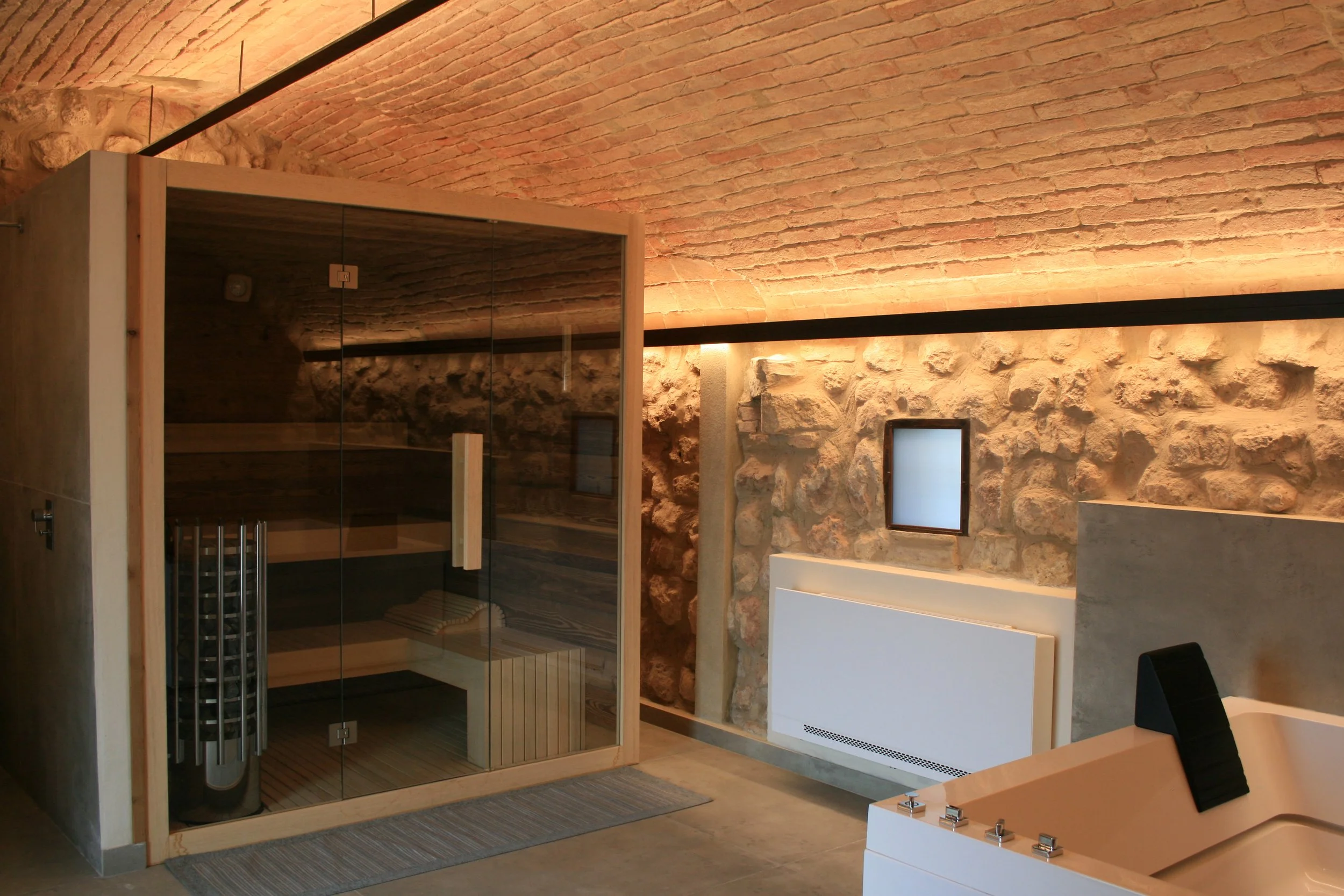 Modern bathroom with exposed brick ceiling and stone wall, featuring a glass-enclosed sauna and a bathtub.