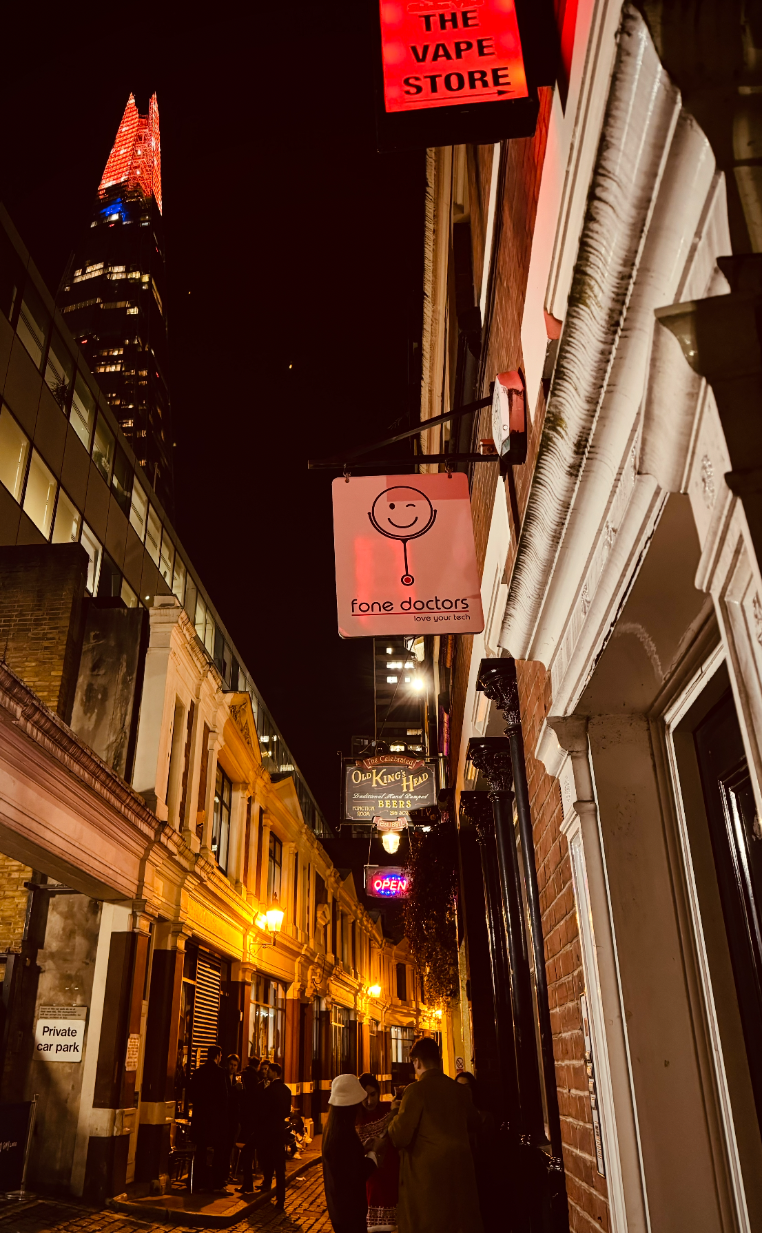 Nighttime street scene showing illuminated storefronts and signs, including a sign for 'Fone Doctors' with a smiling face logo, and other signs reading 'The Vape Store,' 'Old King's Head,' and an 'Open' sign. People are gathered on the sidewalk, some