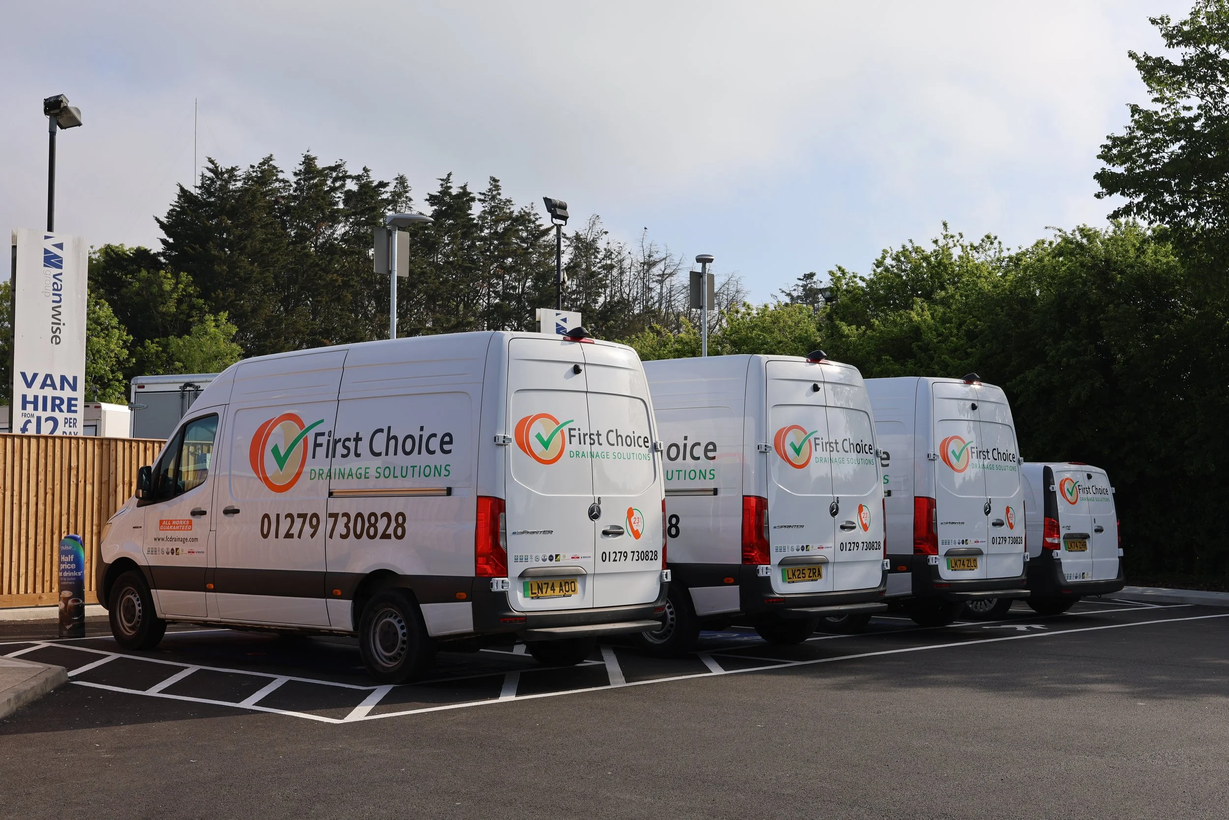 Four white vans with 'First Choice Drainage Solutions' logos and contact information parked in a lot, with trees and a fence in the background.