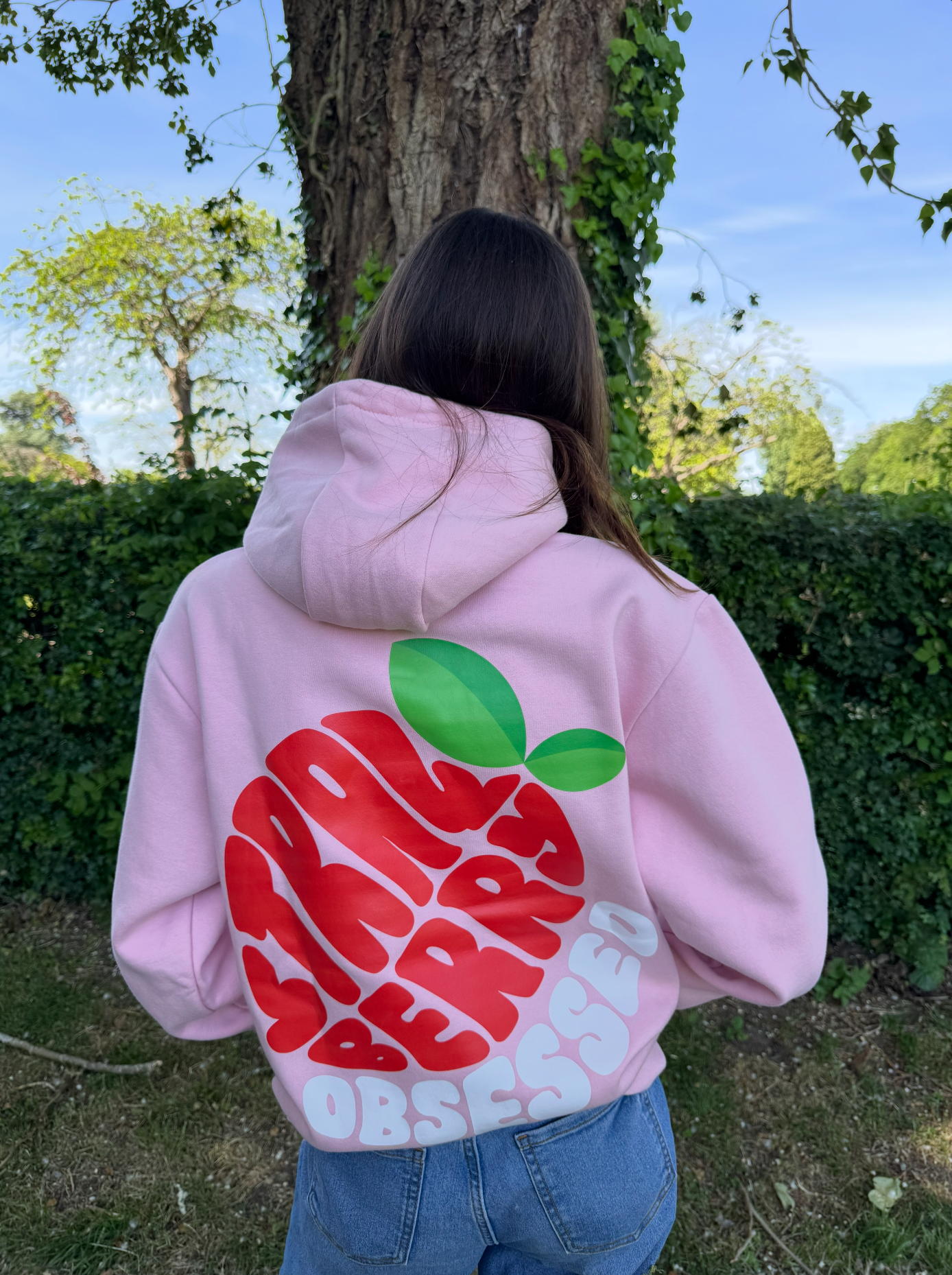 Back of a person wearing a pink hoodie with a red and green graphic and the words "OBSESSED" on the back, standing outside near a tree and greenery.