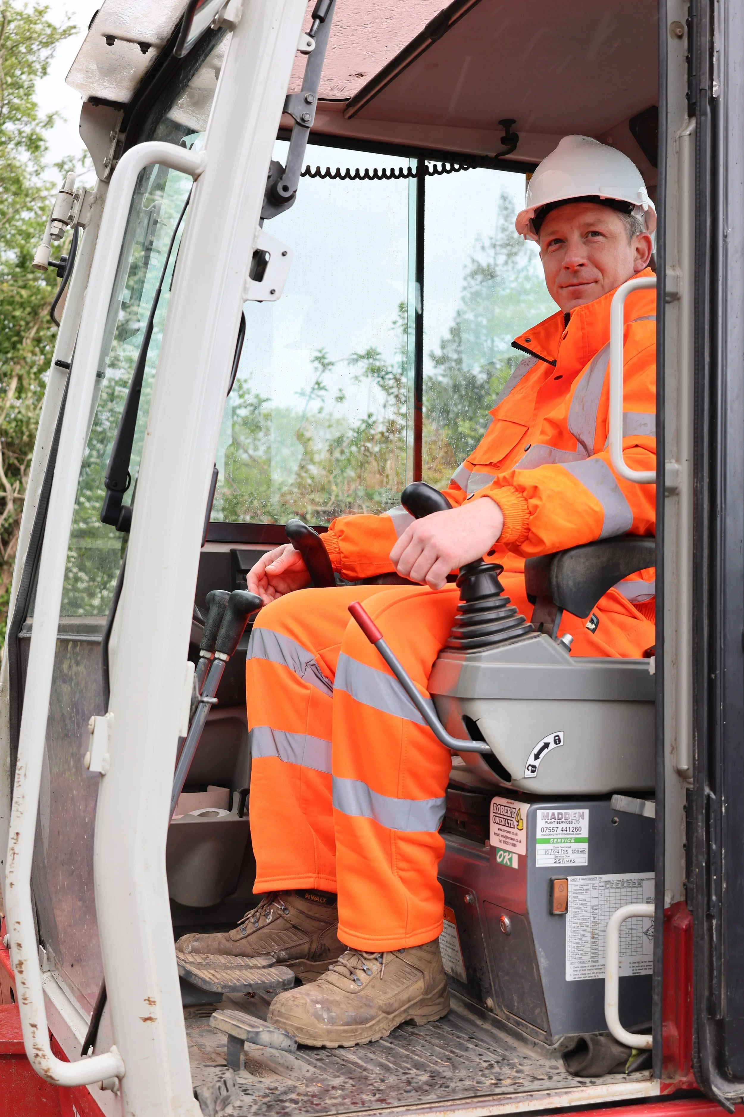 A construction worker in an orange safety suit and white helmet sits in the cabin of a construction vehicle, possibly a bulldozer or excavator, holding the controls.