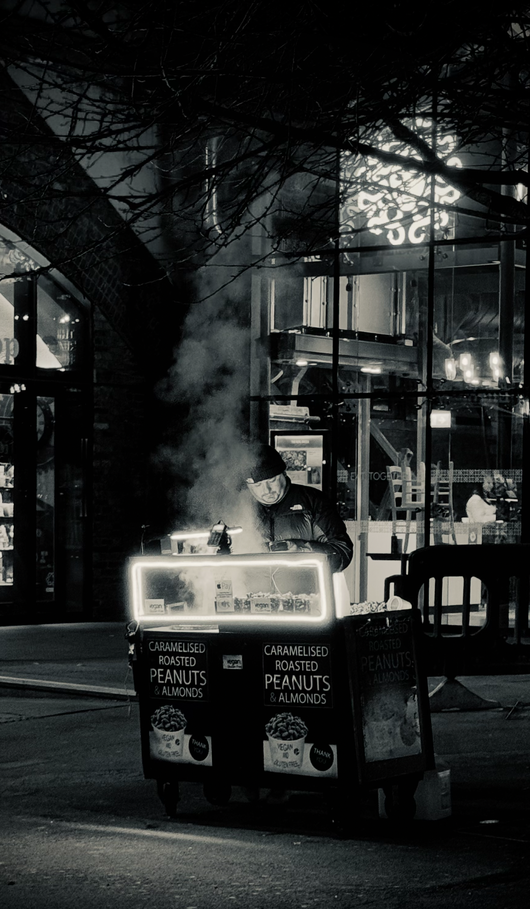 A street vendor selling caramelized roasted peanuts and almonds at night, with steam rising from the cart, illuminated by surrounding streetlights and signage.