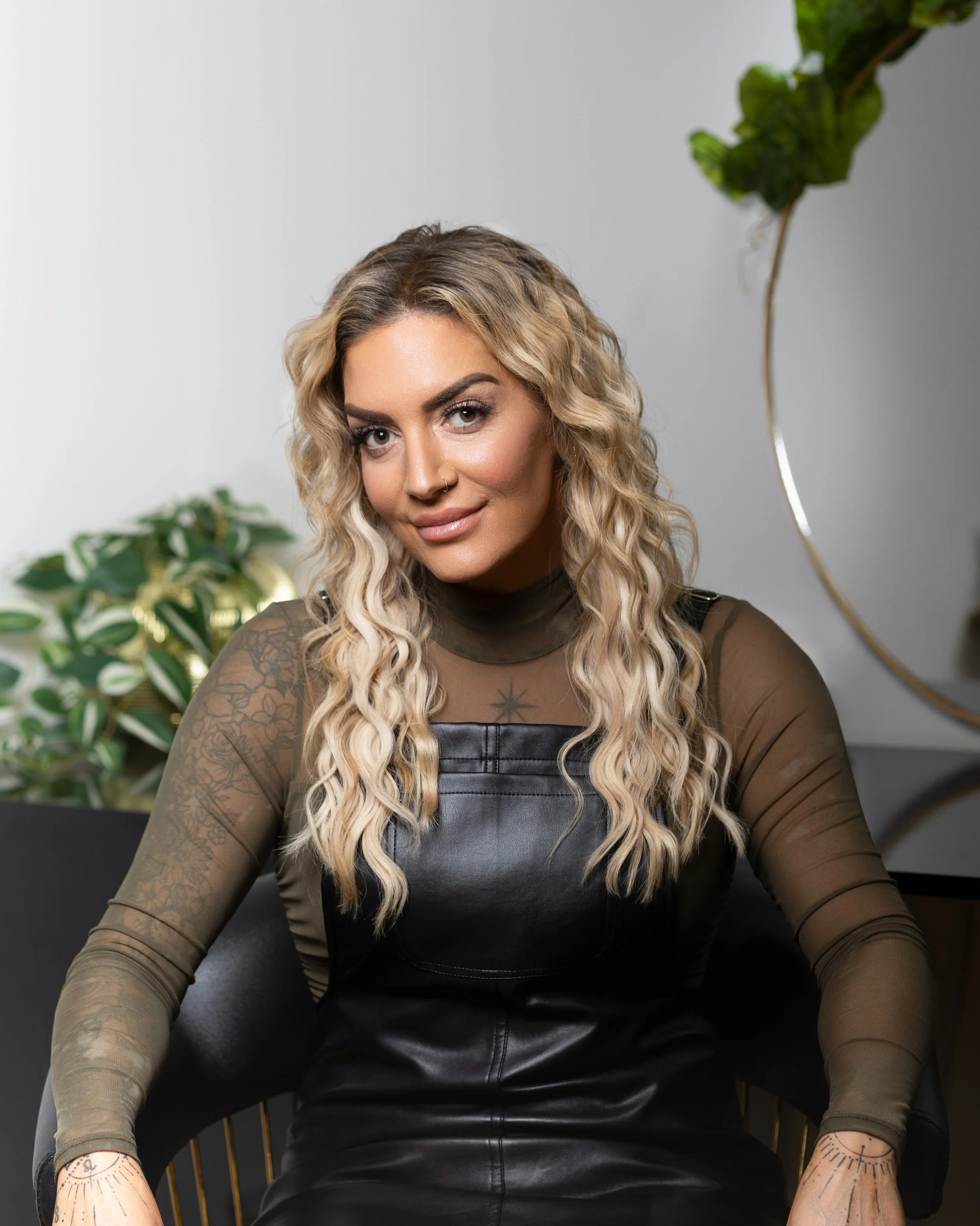 A woman with wavy blonde hair, wearing a see-through olive green long sleeve top and a black leather dress, seated on a modern chair with plants in the background.