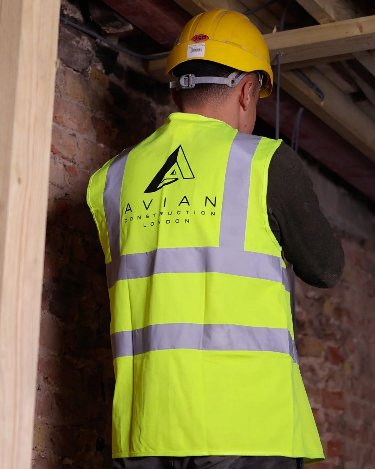 A construction worker wearing a yellow safety helmet and a high-visibility vest with the logo and name "Avian Construction London" printed on the back, working inside a building with brick walls and wooden beams.