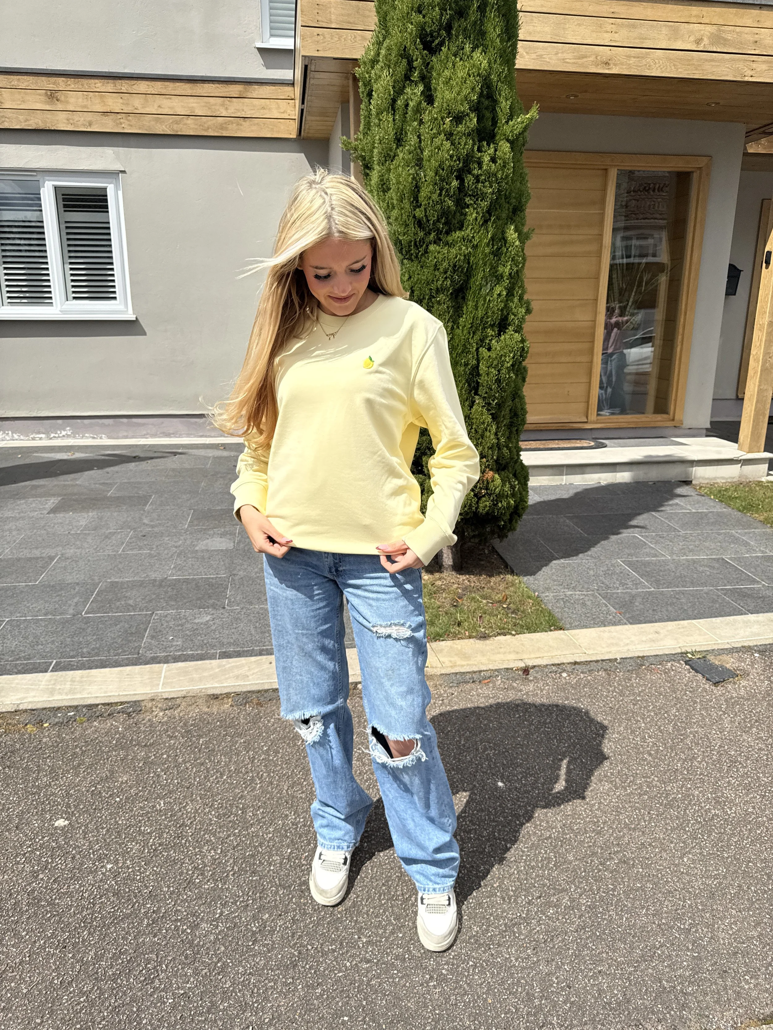 A young woman standing outdoors on a paved sidewalk, wearing a yellow sweatshirt, ripped blue jeans, and white sneakers, with long blonde hair and looking down.