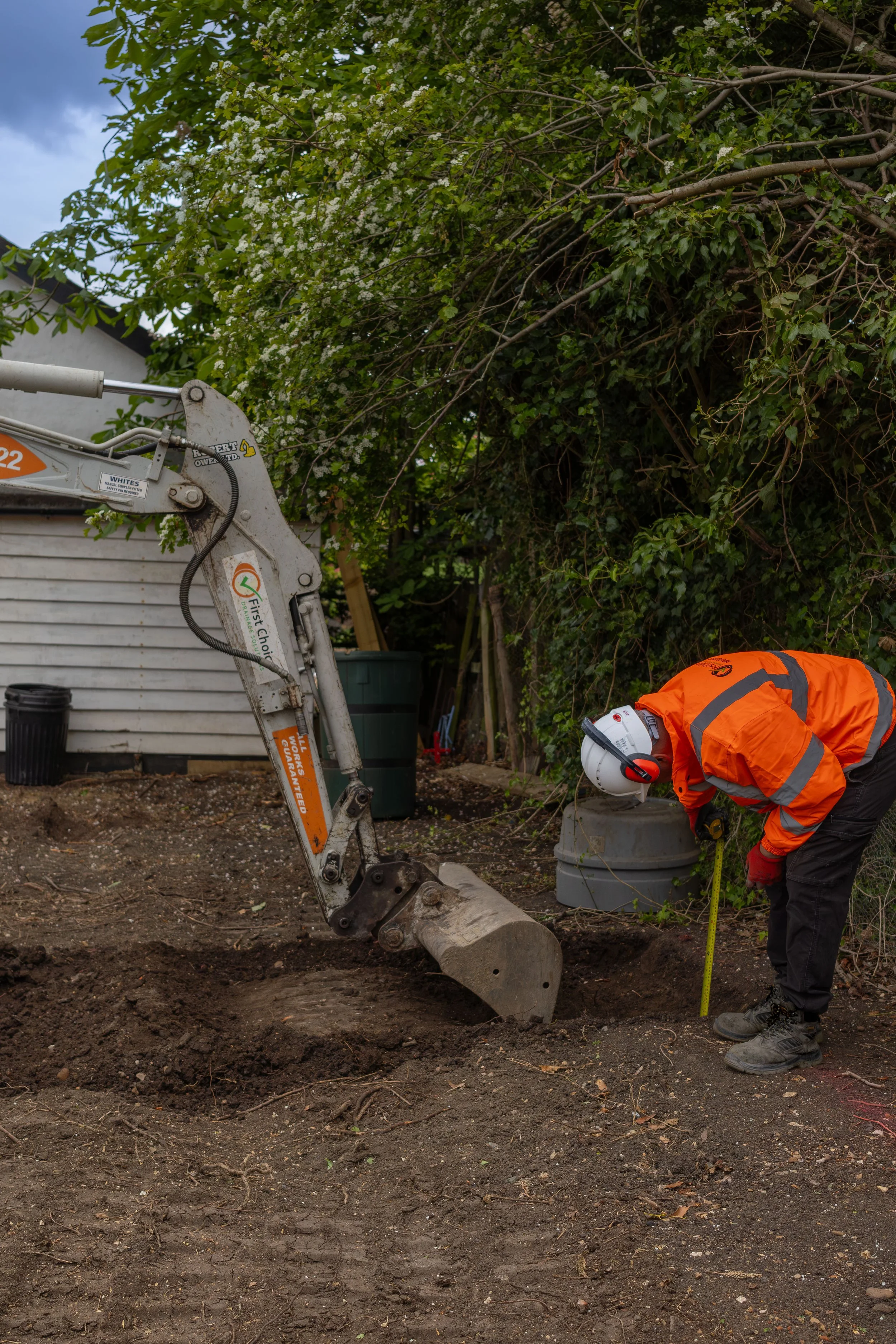 A construction worker wearing orange safety gear, including a high-visibility jacket and a helmet with ear protection, is working on a construction site next to a small excavator. The worker is holding a measuring tape and appears to be measuring the