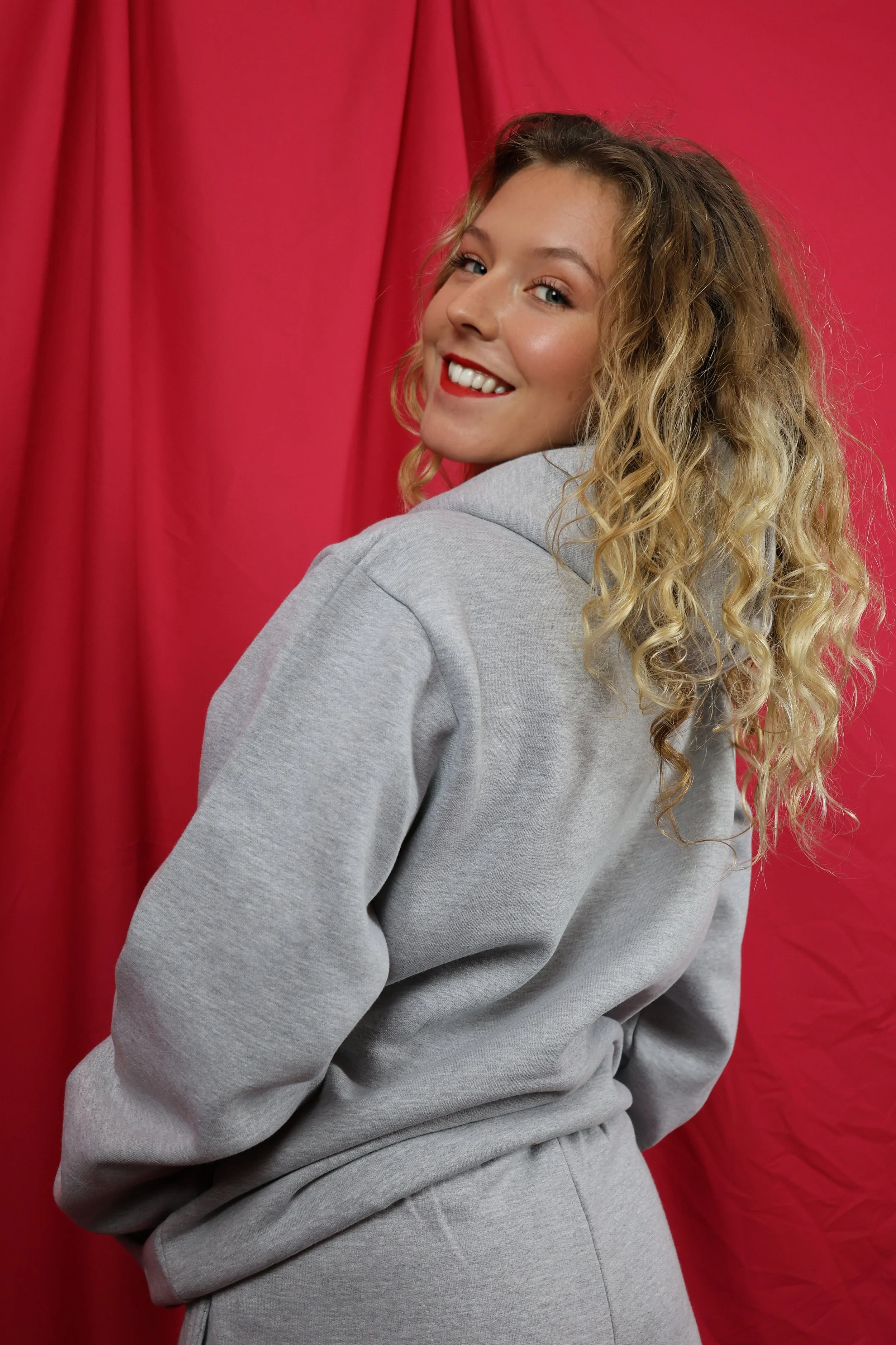 A young blonde woman with curly hair smiling and looking over her shoulder, wearing a gray hoodie and sweatpants in front of a red curtain backdrop.
