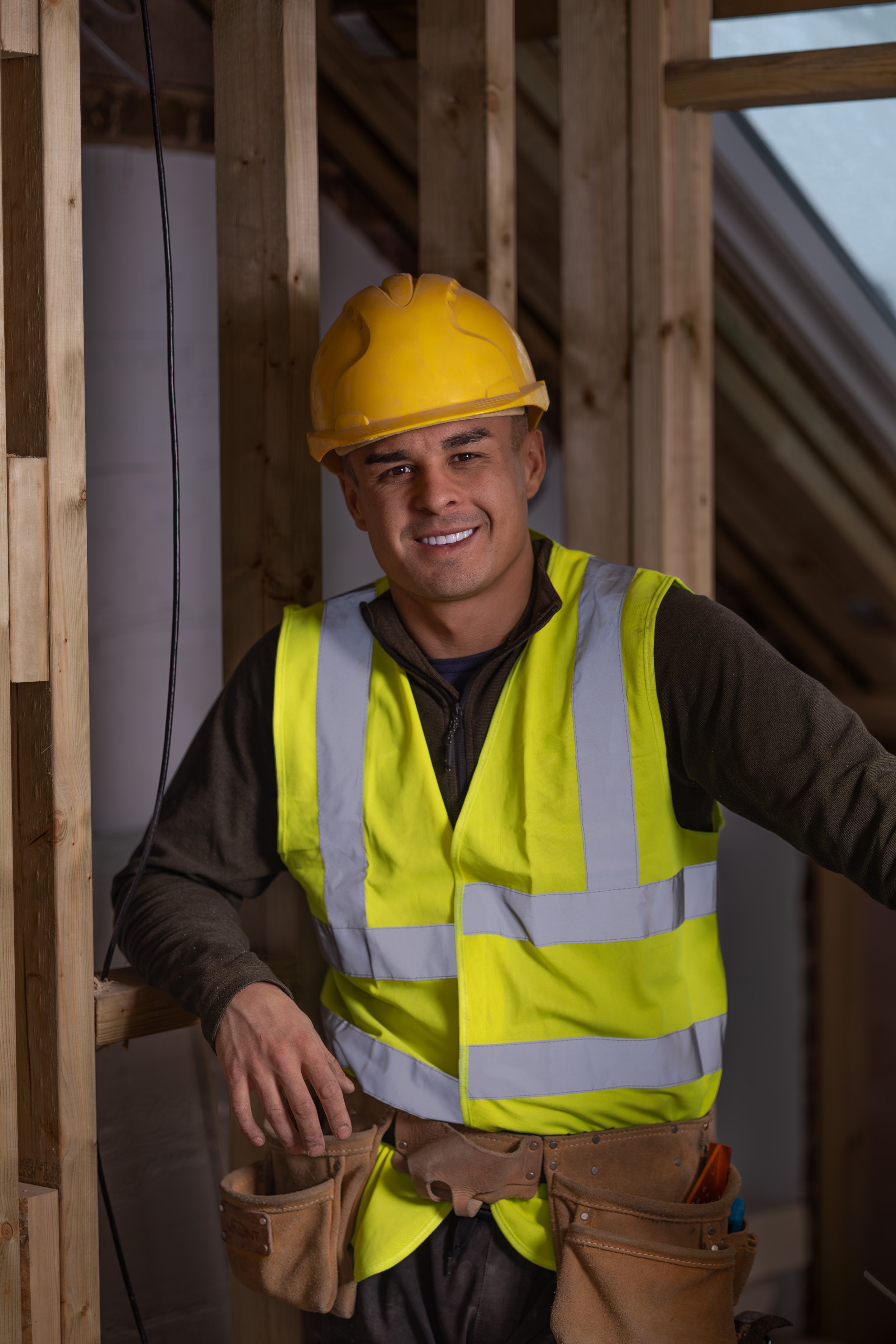 A smiling construction worker wearing a yellow hard hat and a neon yellow safety vest inside a wooden frame of a building under construction.
