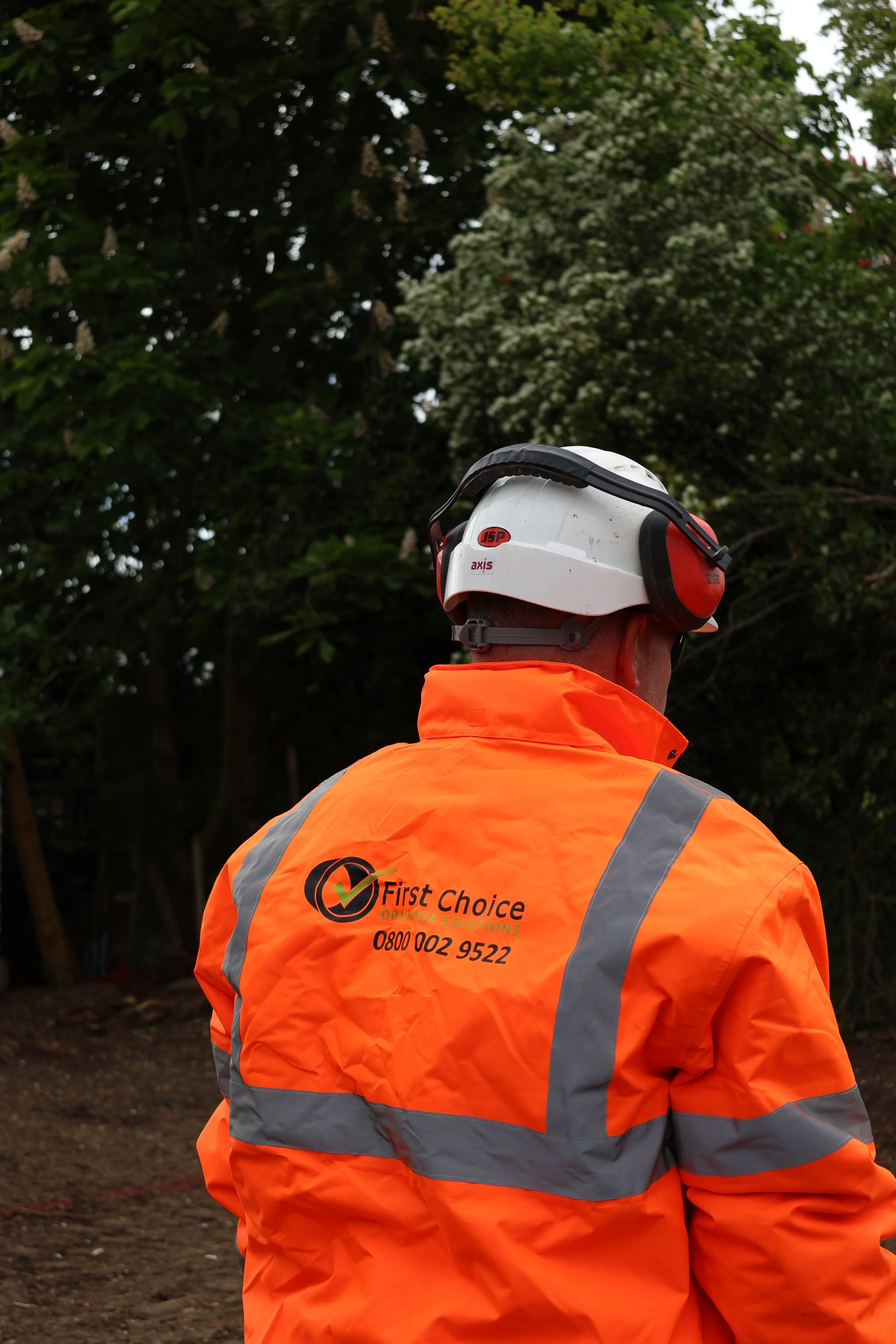 A person wearing an orange safety jacket and white helmet with headphones, with their back facing the camera, standing outdoors.