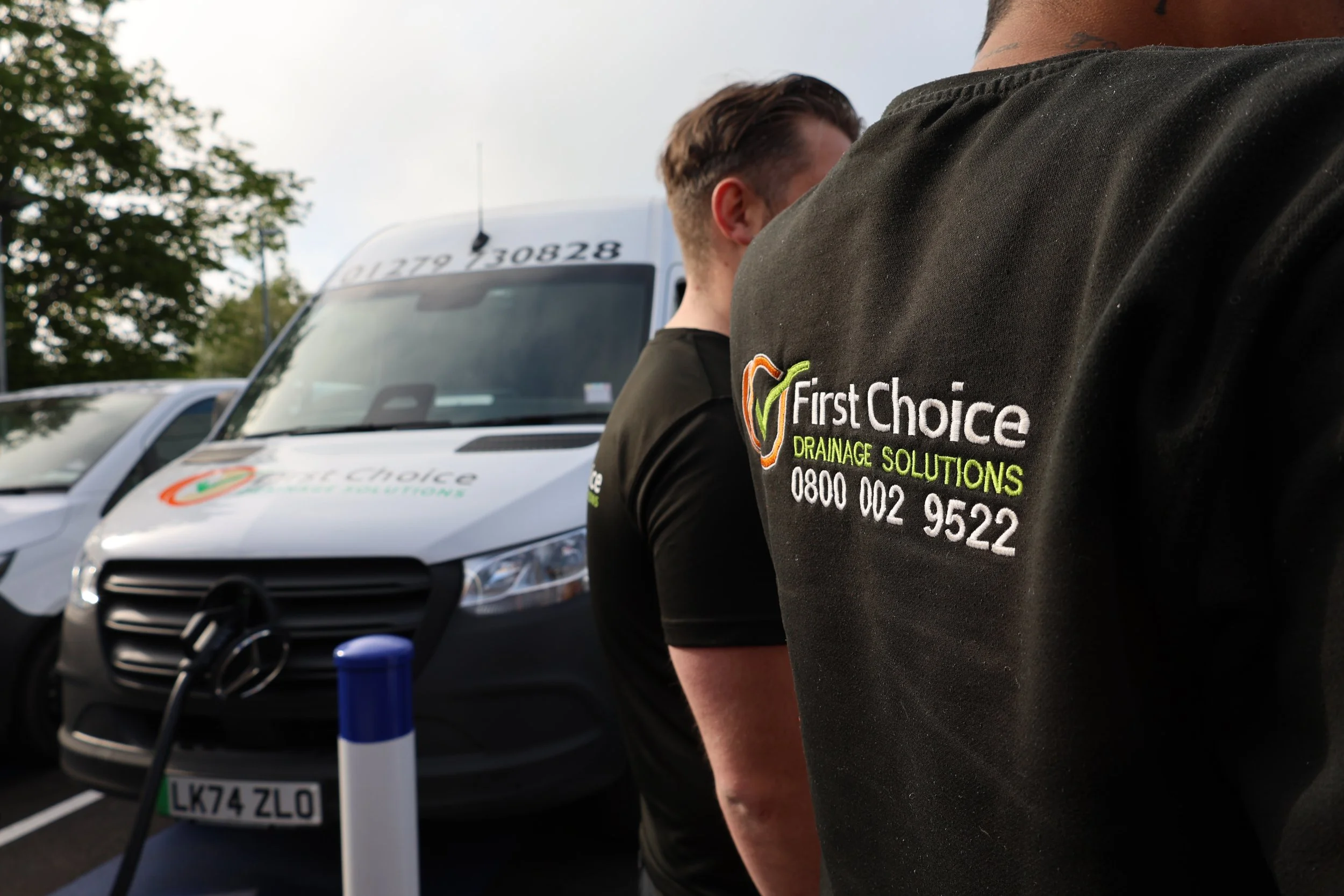 Close-up of two workers wearing black shirts with 'First Choice Drainage Solutions' logo, standing near a company van with the same logo, in a parking lot.
