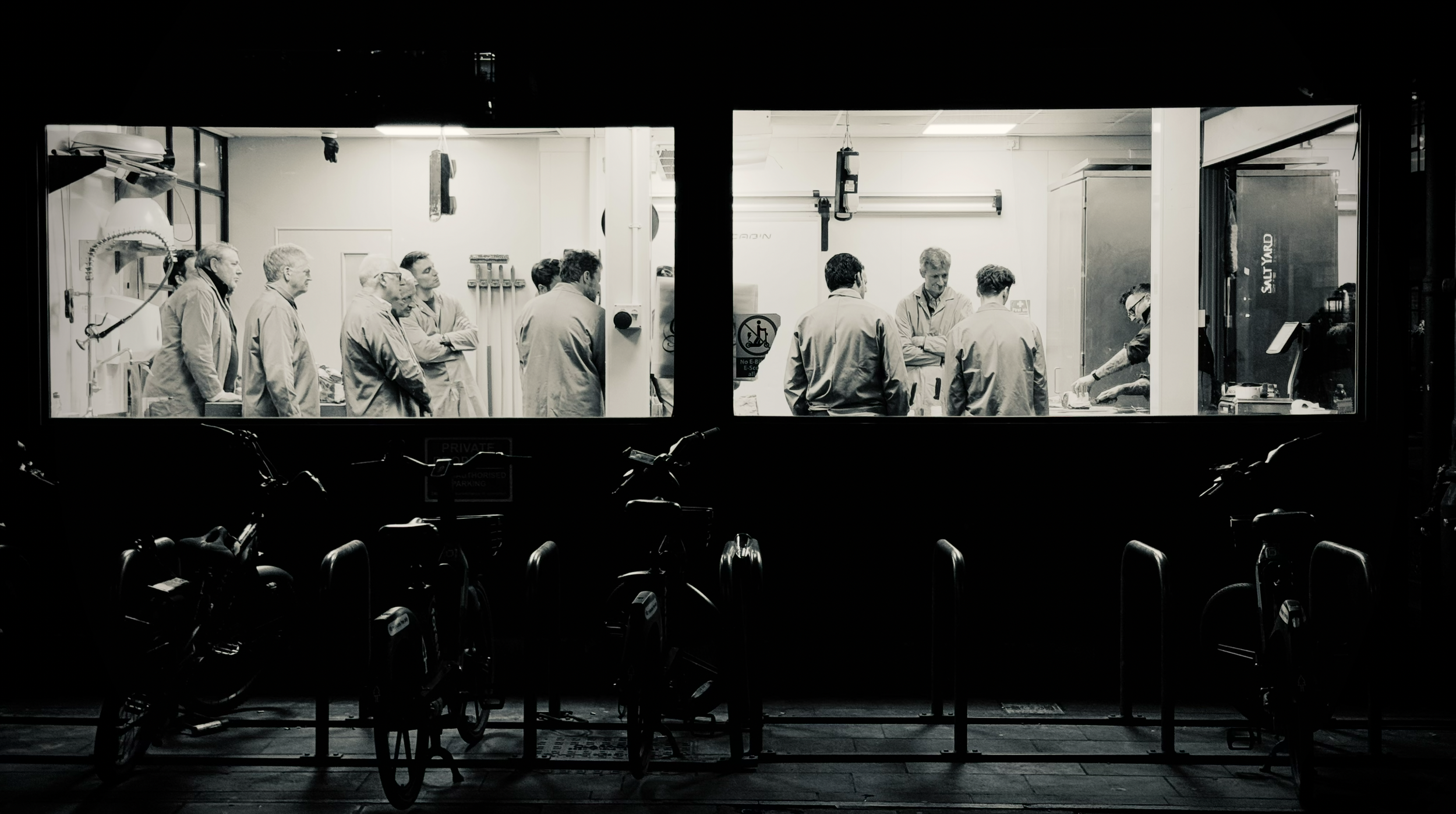 A black and white photo of people gathered inside a room, seen through a large window. Bicycles are parked outside in front of the window.