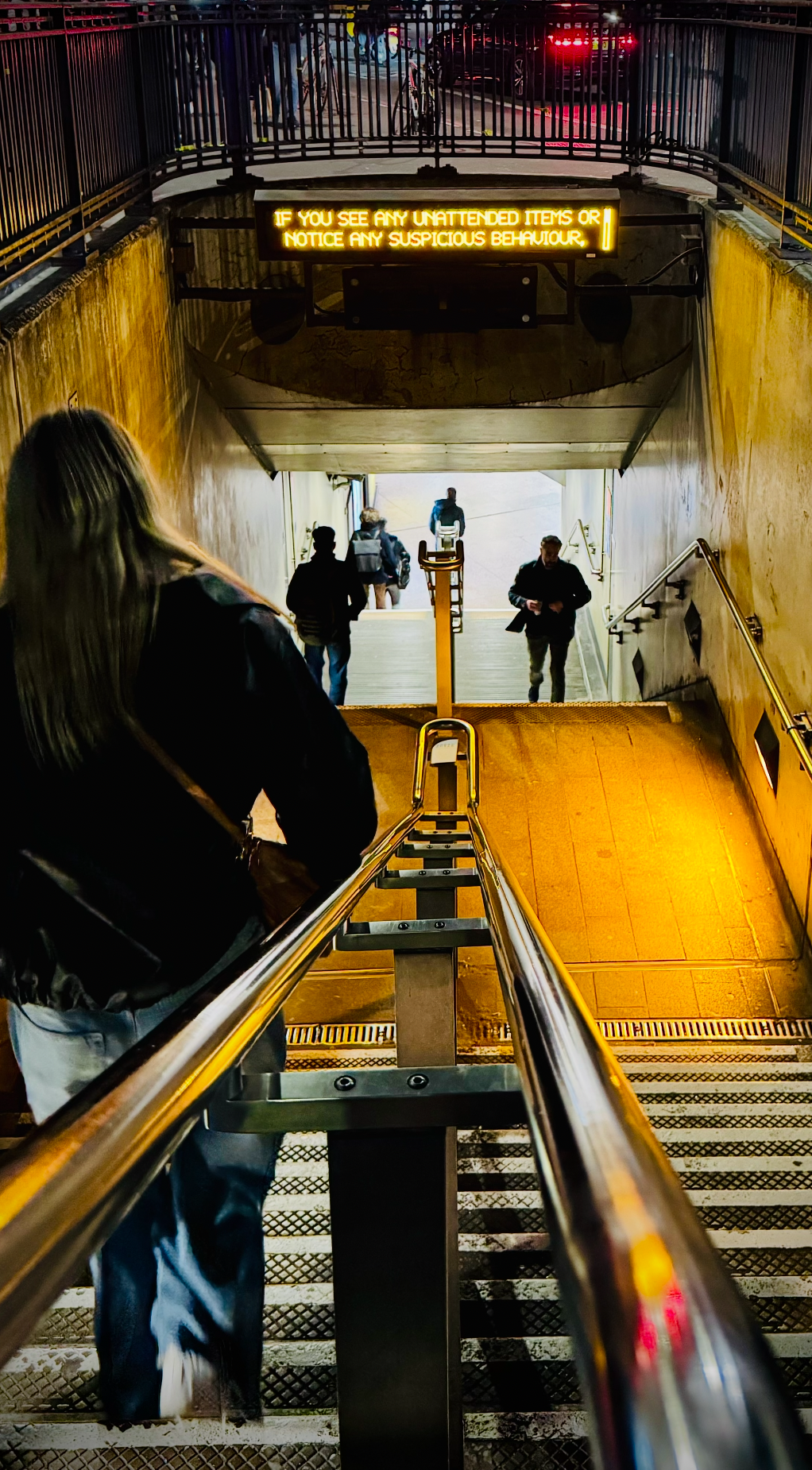 People descending underground through a stairway in a subway station, with a yellow wall and a digital sign warning about suspicious behavior.