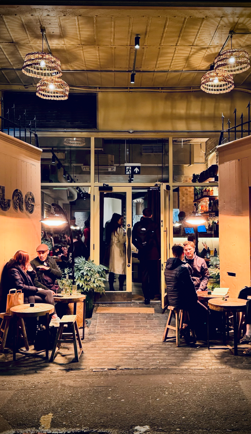 Nighttime scene outside a cozy café or restaurant with people sitting at small tables, talking and enjoying drinks. Inside, more people are seen through glass doors, with warm lighting and a bar area in the background.