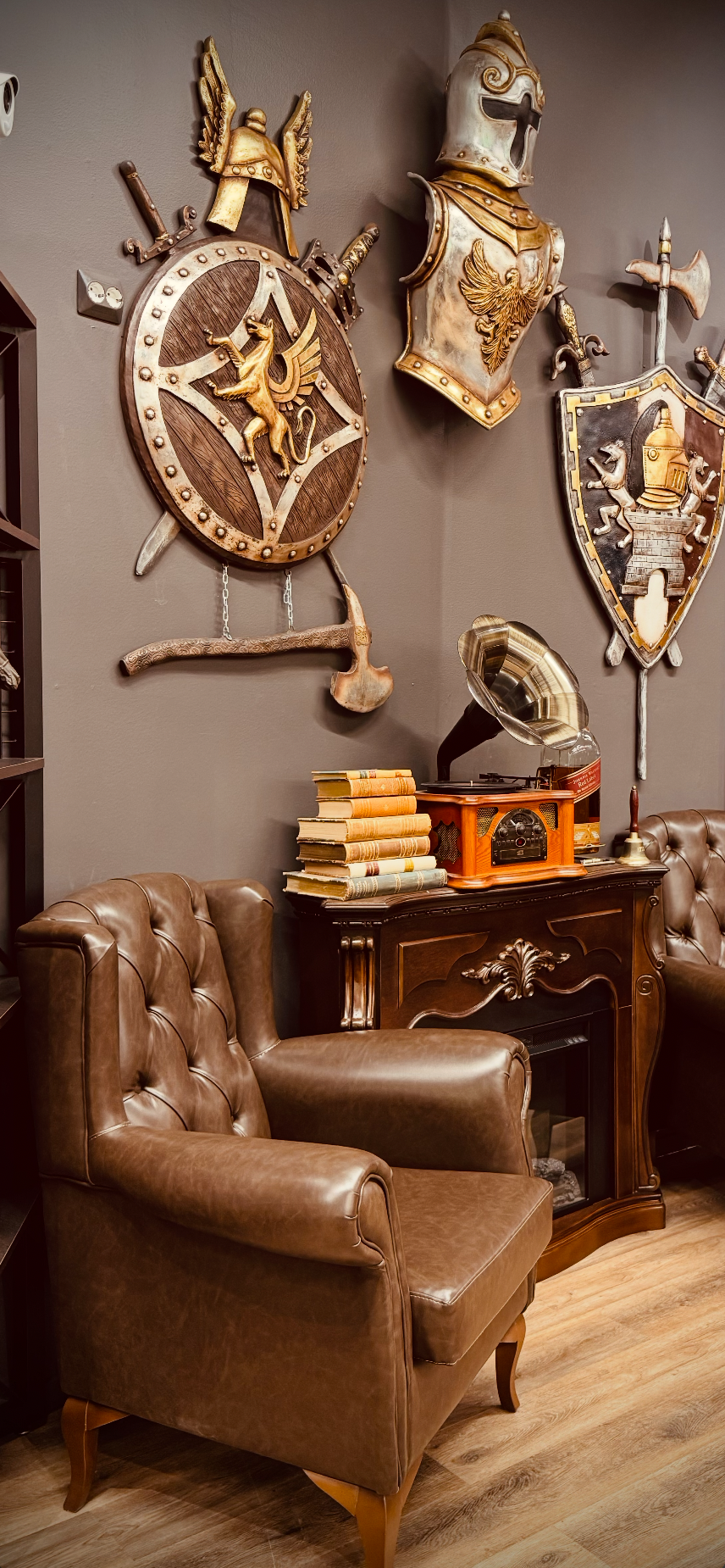 A wall decorated with medieval armor and shields, including a helmet, a coat of arms, and various swords, above a vintage wooden cabinet with a stack of old books and a gramophone, in a cozy living room setting.