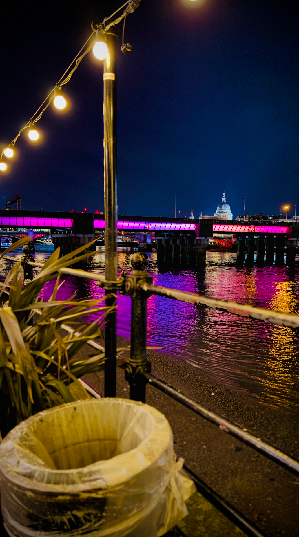 Night scene of the River Thames in London with a bridge lit in pink and purple lights, the dome of St. Paul's Cathedral in the background, a metal railing, string lights overhead, and a trash bin in the foreground.