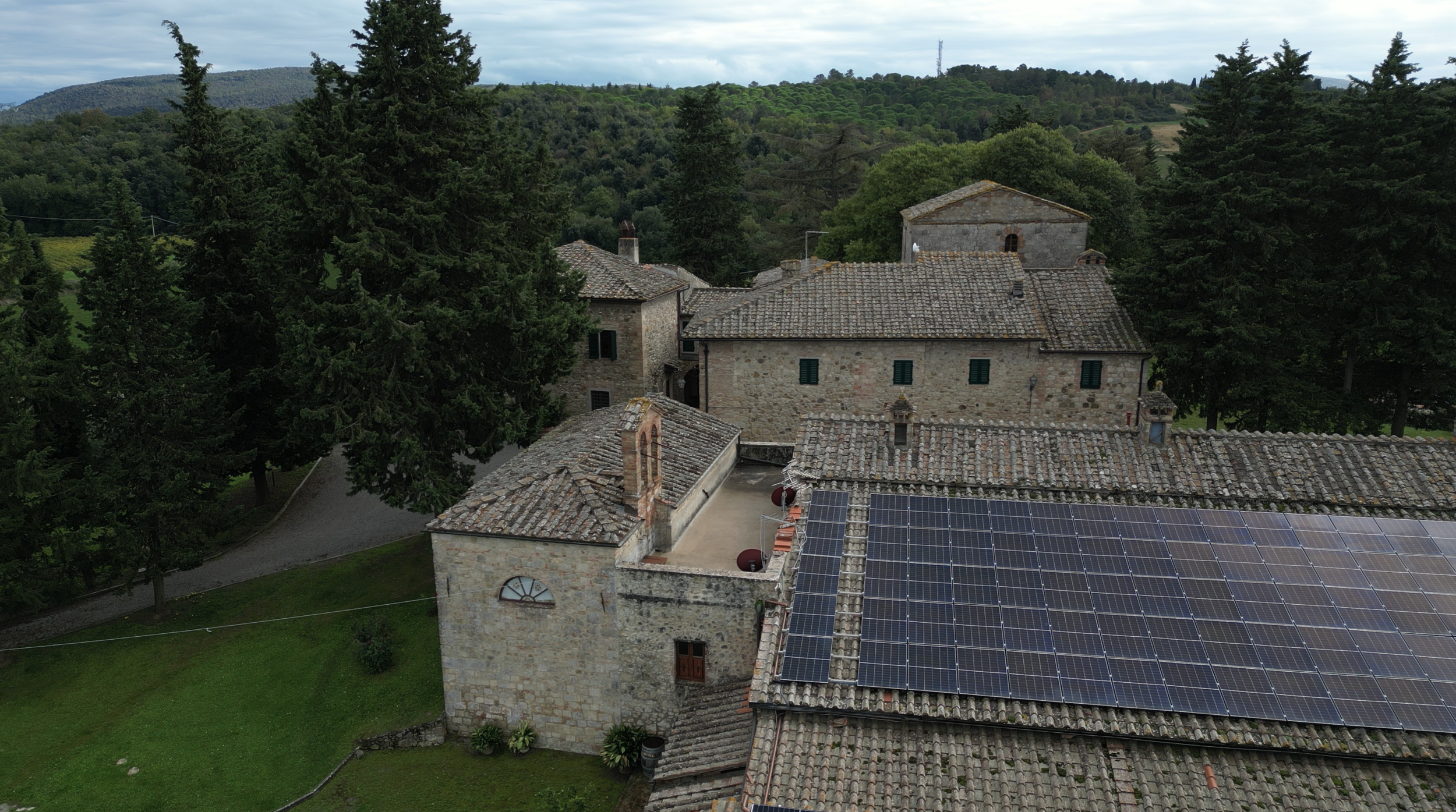 A rustic hillside village surrounded by lush green trees, with old stone buildings and tiled roofs, some equipped with solar panels.