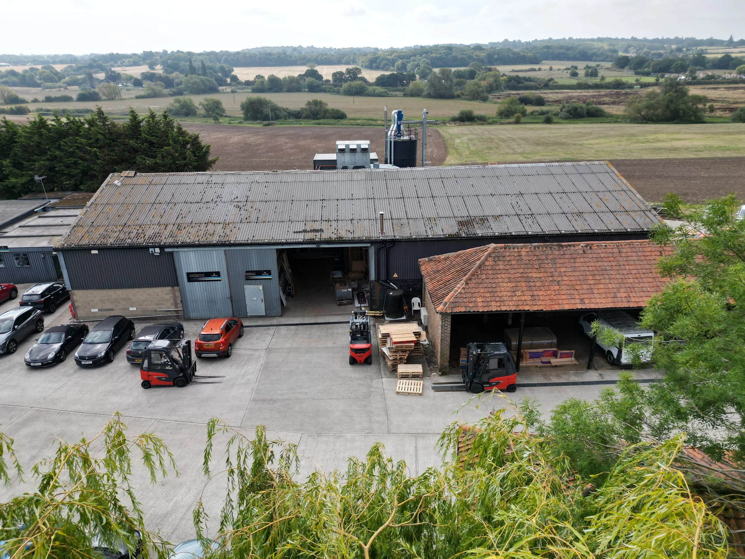 Aerial view of a warehouse with a metal roof, parked cars, and forklifts, surrounded by farmland and greenery.