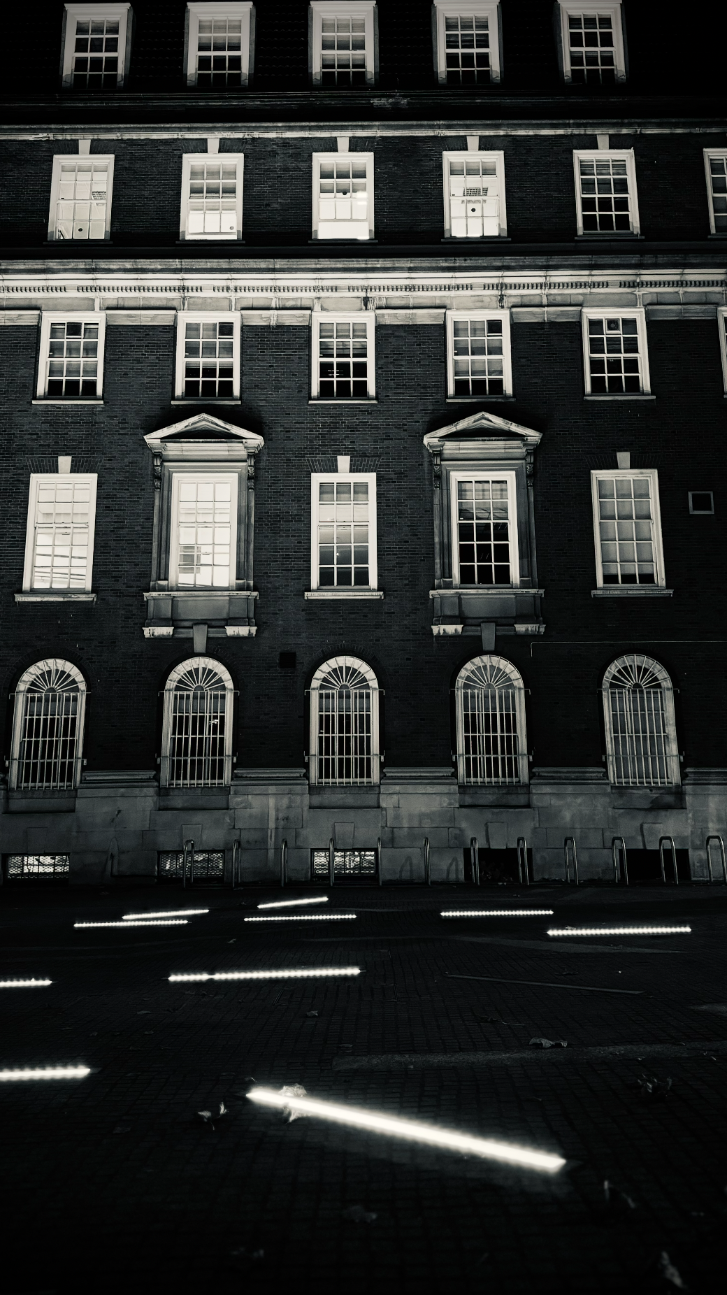 Nighttime view of a tall brick building with multiple windows, some with bars, and decorative architectural details, illuminated by streetlights and streaks of light on the ground.