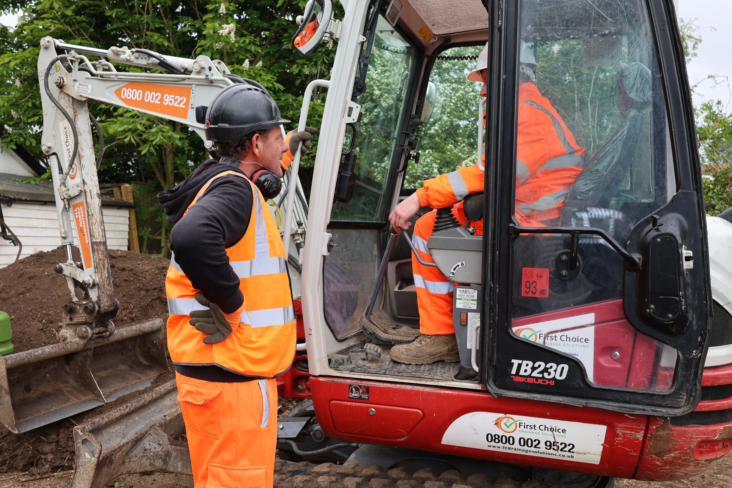 Two construction workers wearing orange safety vests, helmets, and gloves, with one sitting inside a small excavator and the other standing outside, having a conversation on a construction site.