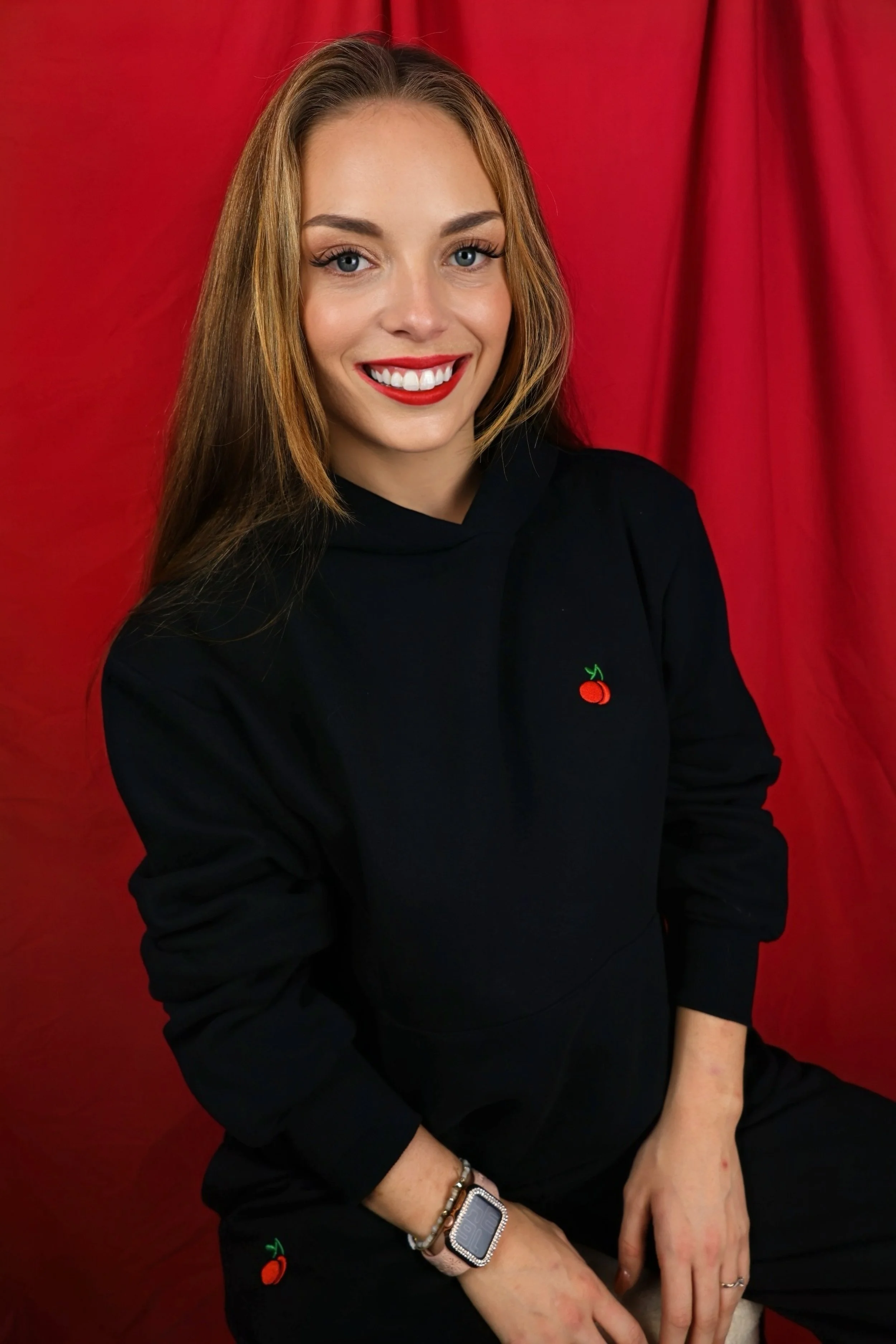 A young woman with long, wavy, light brown hair, blue eyes, wearing red lipstick, a black hoodie with a small cherry embroidered on the chest, sitting against a red curtain backdrop, with a smartwatch and jewelry on her wrist.