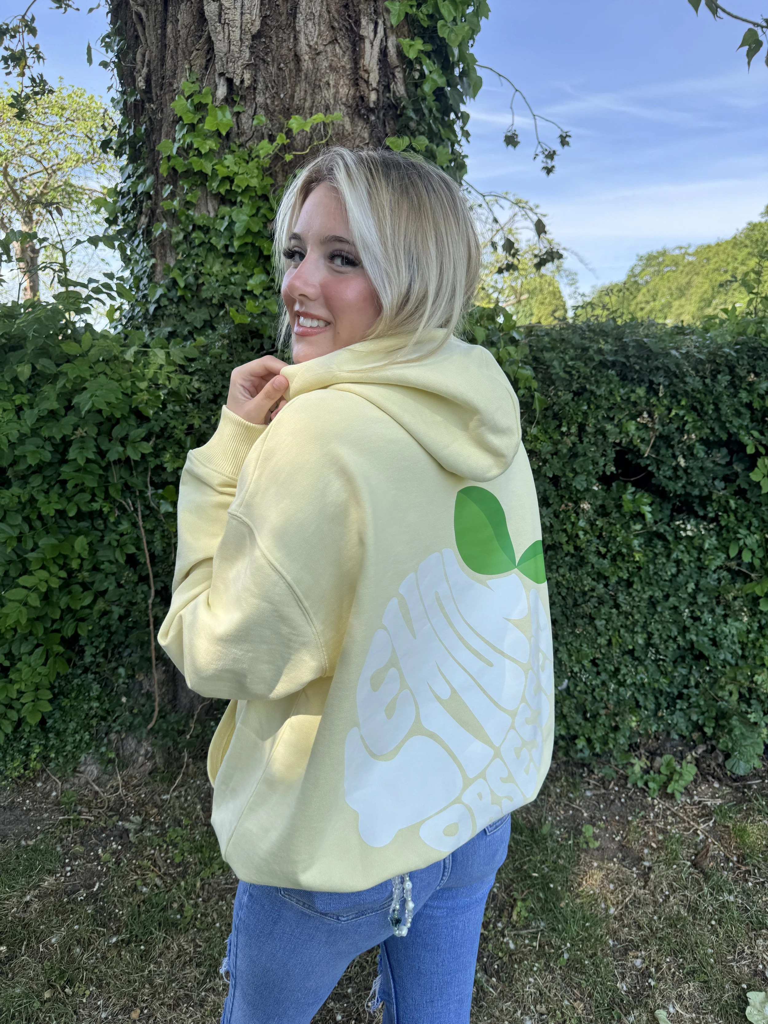 A young woman with blonde hair outdoors, wearing a yellow hoodie with a large white and green design on the back, standing in front of a tree and green foliage.