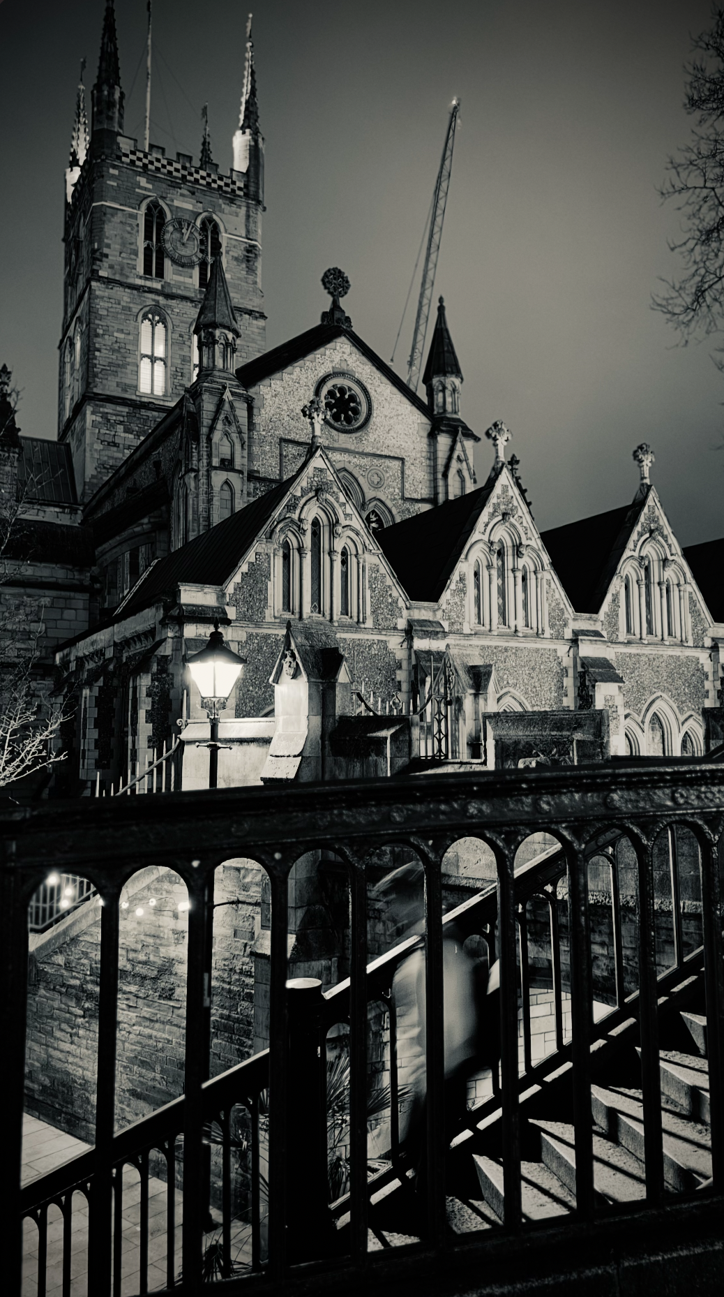 Nighttime view of a historic church with Gothic architecture, tall spires, arched windows, and a clock on the tower, seen through a metal fence.
