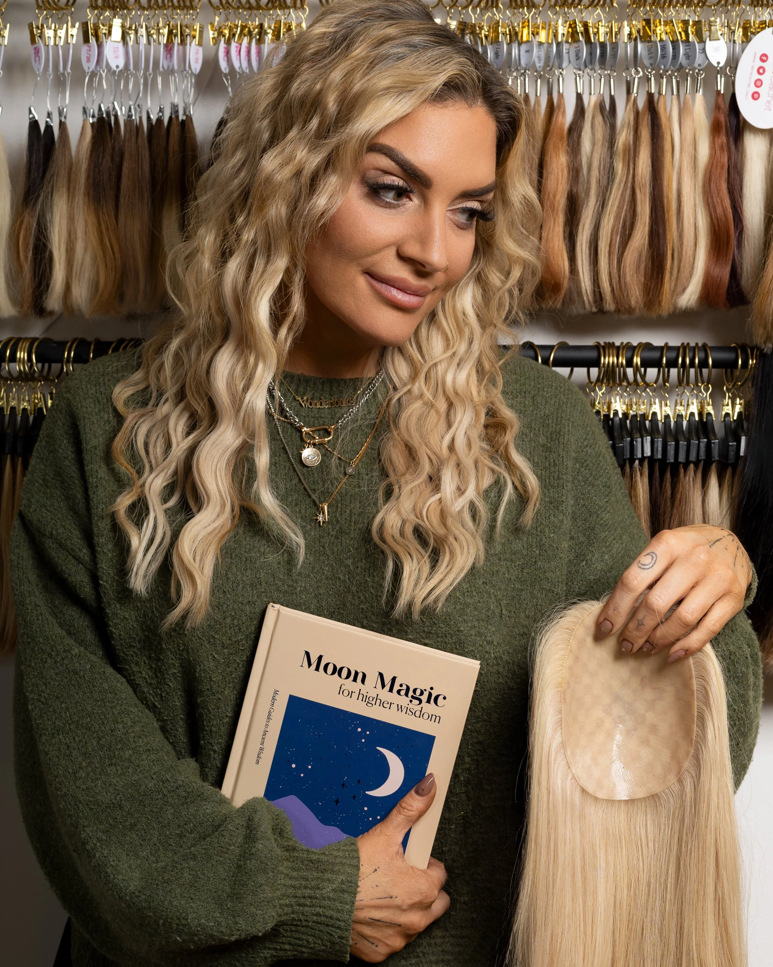 A woman with long, curly blonde hair shopping for hair extensions in a store, holding a book titled 'Moon Magic for higher wisdom' and examining a blonde hair extension piece.