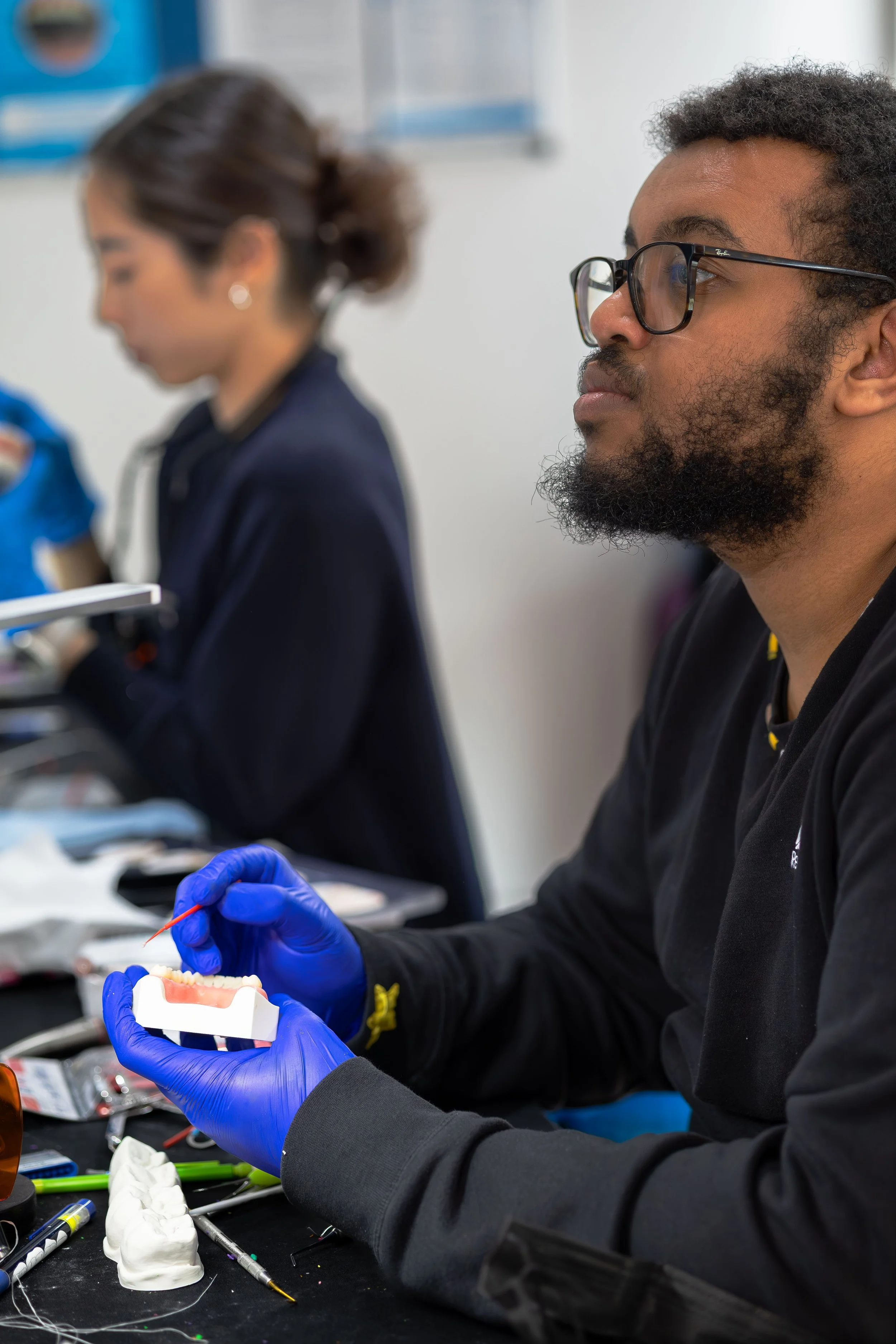 A man wearing glasses and a black jacket working with dental prosthesis or model, using tools and wearing blue gloves, with dental tools and materials on the table.