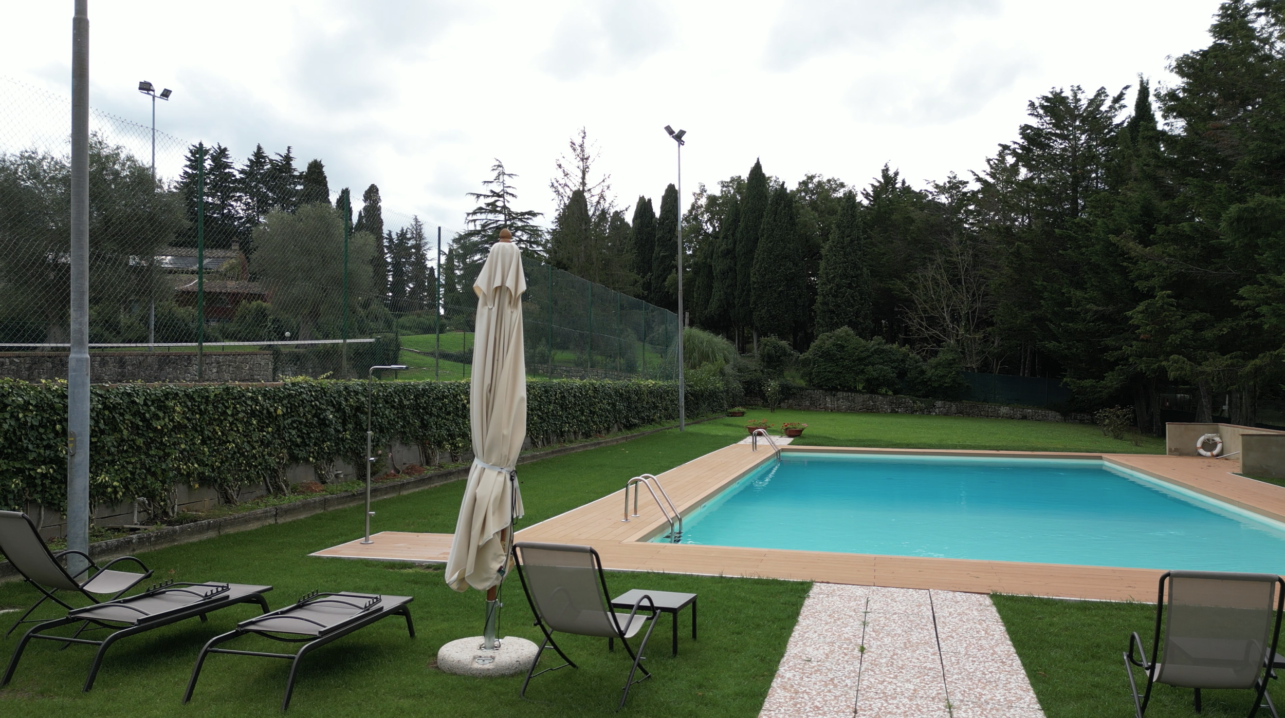 Empty swimming pool with lounge chairs and an umbrella on a grassy area surrounded by trees and a fence under a cloudy sky.