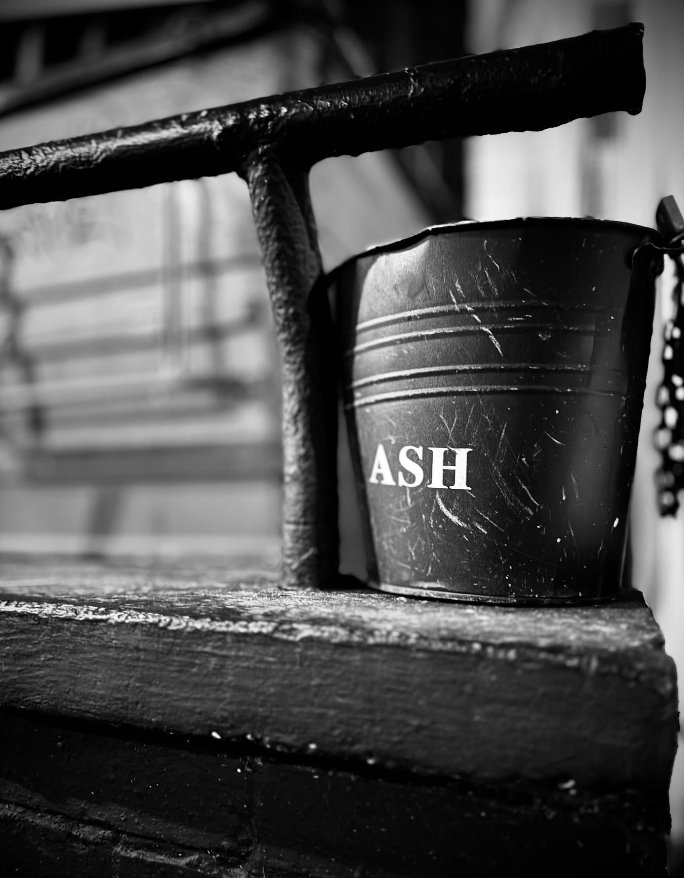 A black plastic bucket with the word 'ASH' written on it in white, placed on a rustic wooden surface and partly obscured by an iron railing.