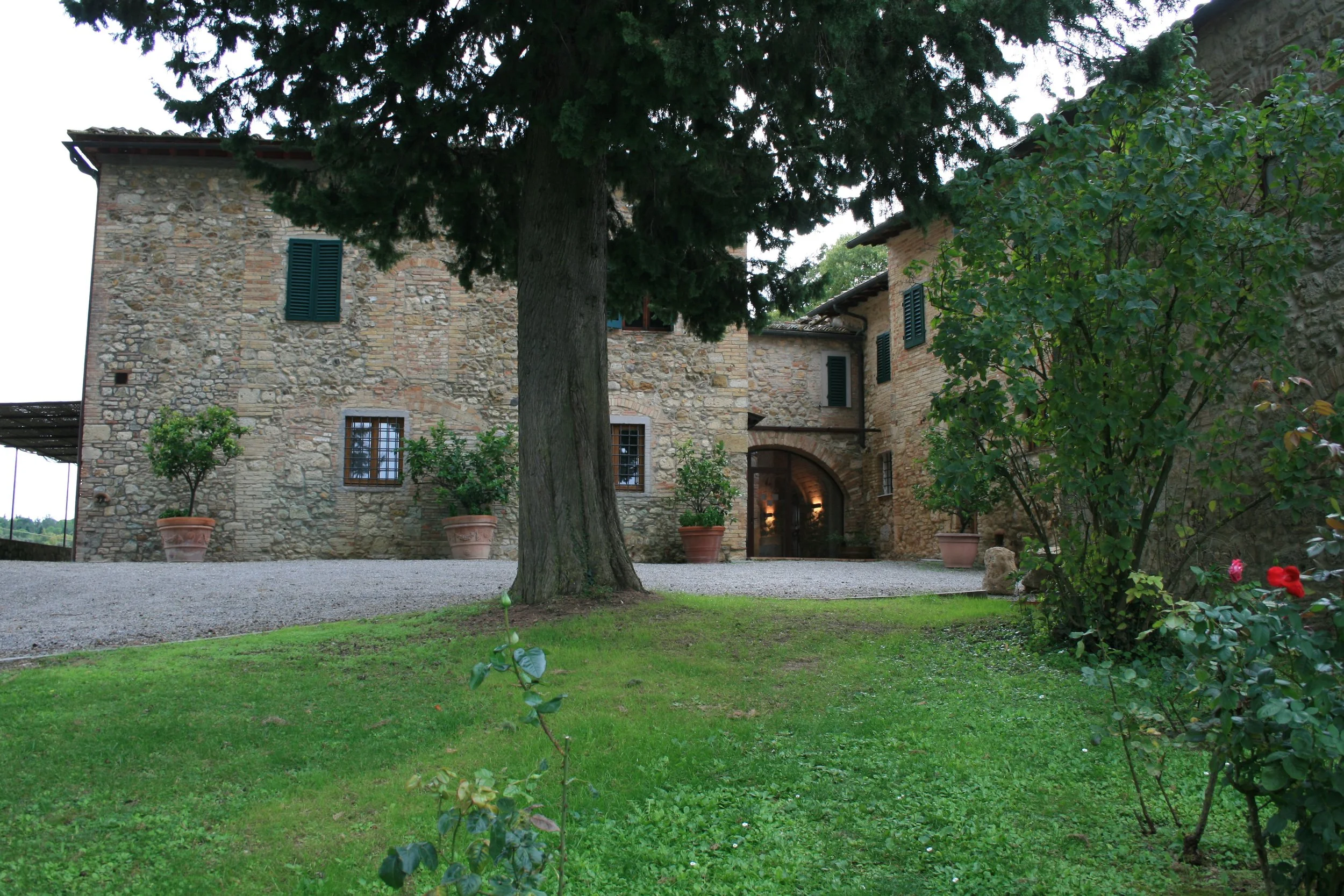 Rustic stone building with green shutters, surrounded by potted plants and trees, and a well-kept lawn.