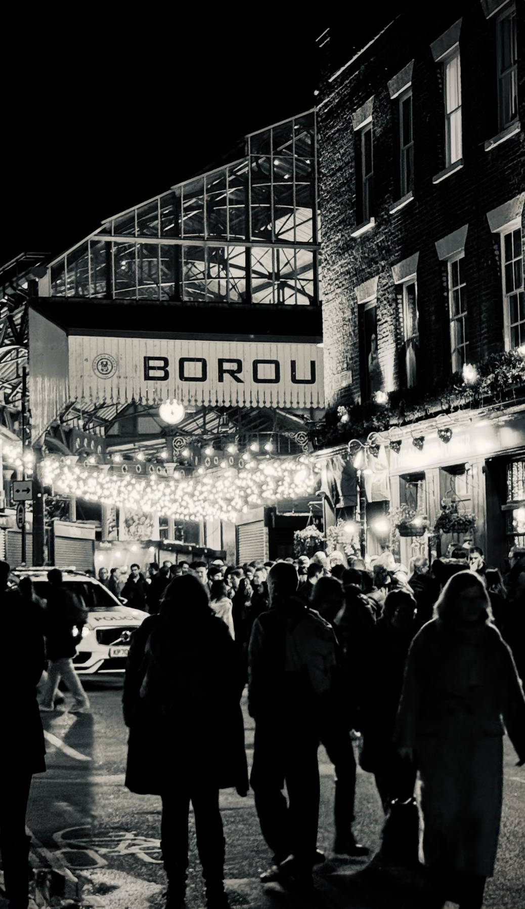Night scene of a crowded street outside a venue called Borou, decorated with string lights and people walking and standing, with a police car visible in the background.