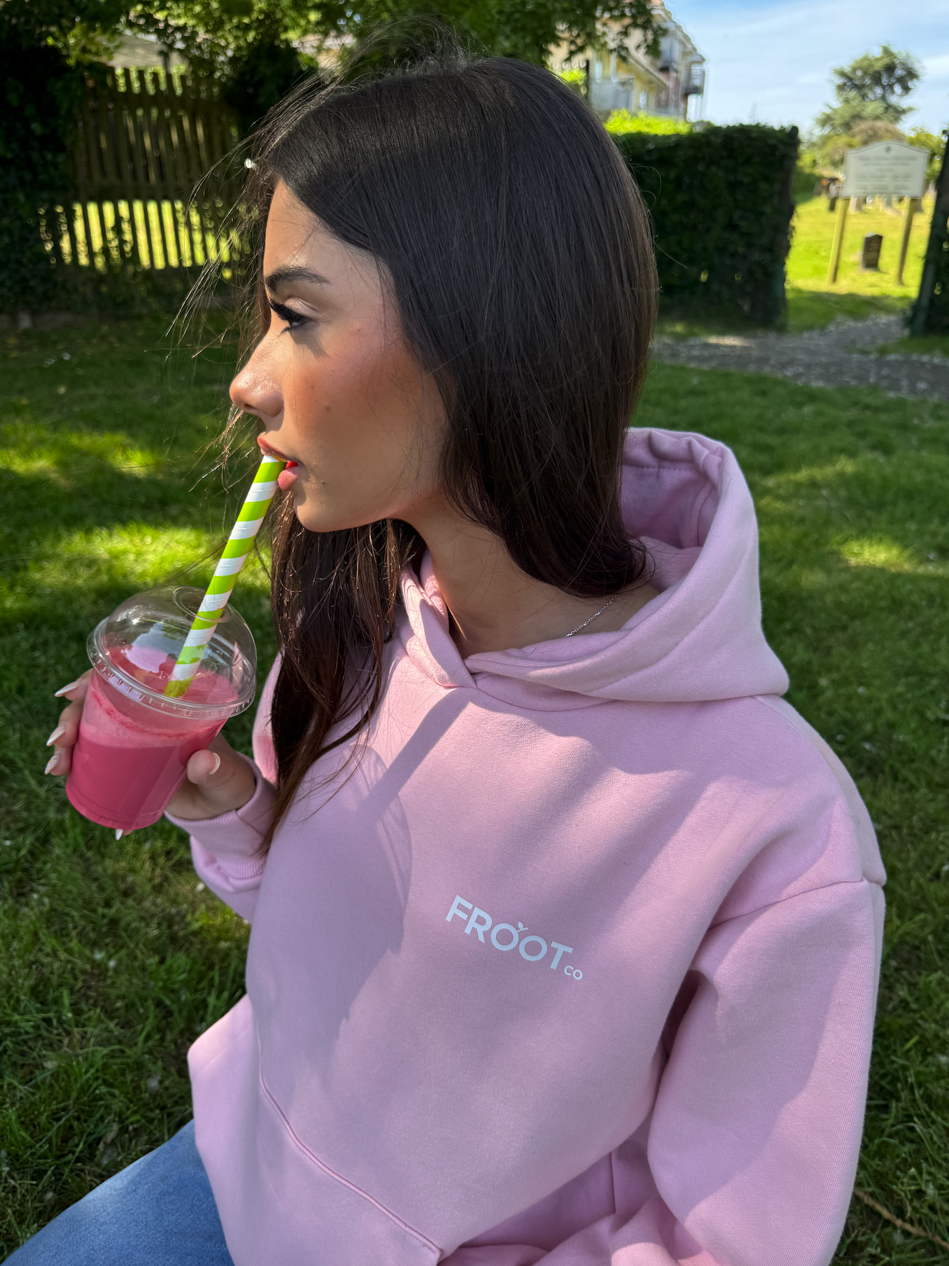 A woman with dark hair wearing a pink hoodie with 'FROOT CO' written on it, drinking a pink smoothie through a striped straw outdoors on a sunny day.