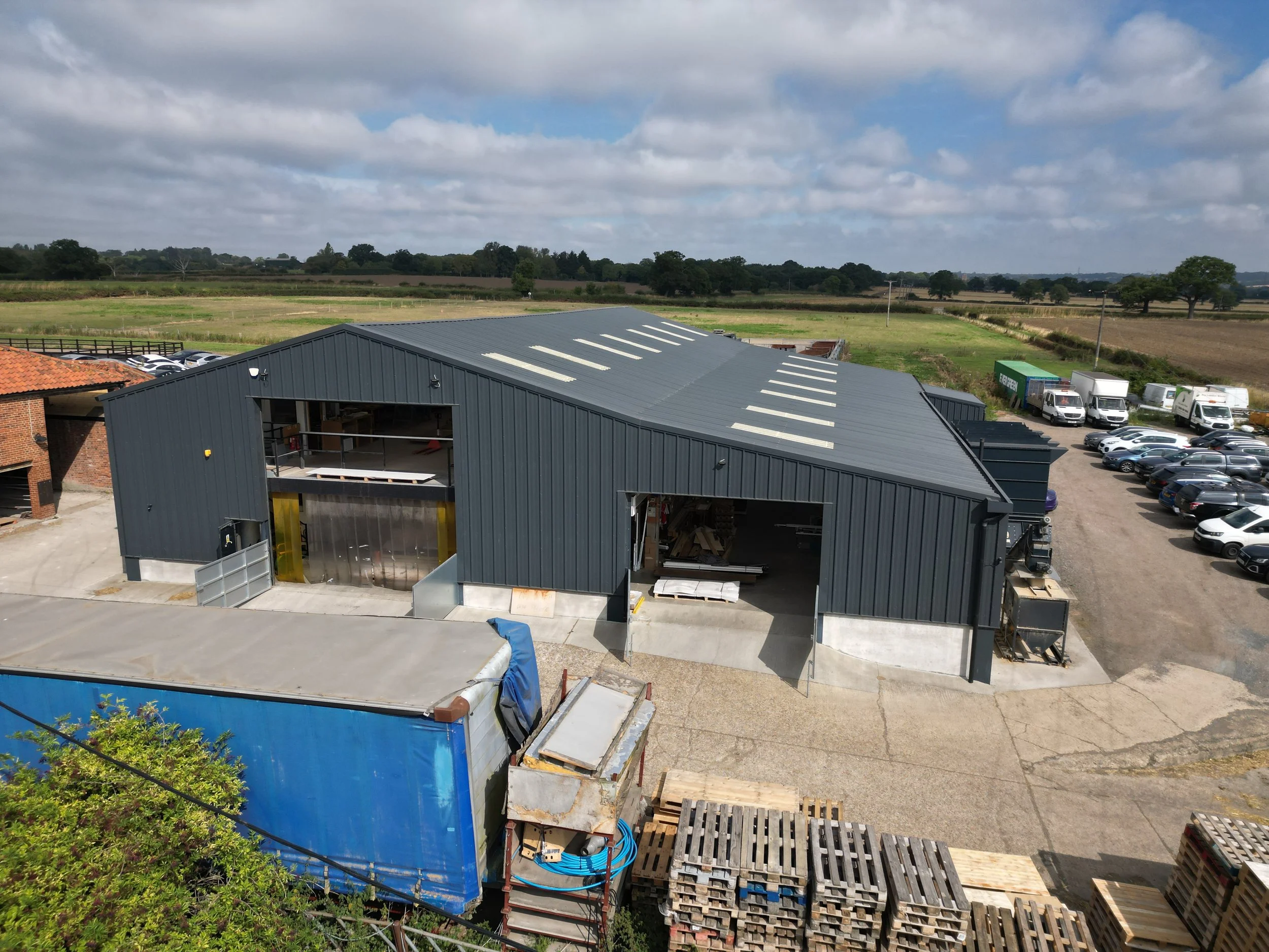 An industrial building with a dark metal exterior under a partly cloudy sky, surrounded by a parking lot with cars, a field, and a clear sky with scattered clouds.