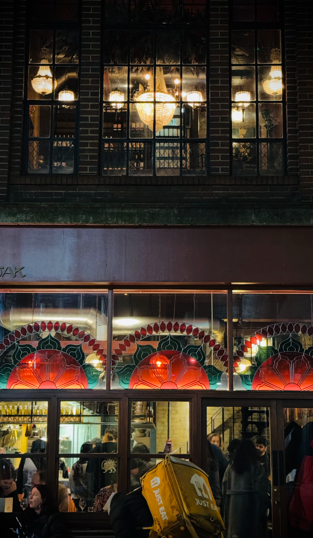 Nighttime view of a restaurant with three large windows, decorated with red and green stained glass panels shaped like flowers or fans. Inside, warm lighting and chandeliers are visible. Outside, people are gathered, some standing and some sitting at