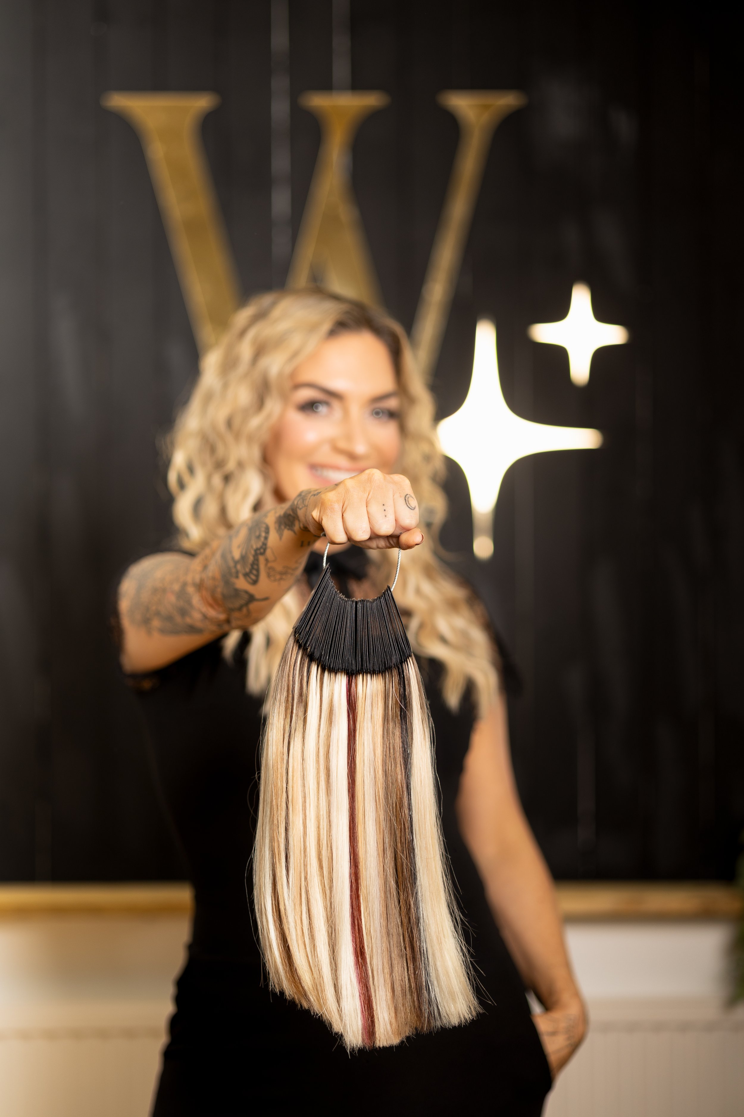 A woman with blonde curly hair holding a hair extension sample in front of a black wall with a gold letter 'W' and star-shaped light.
