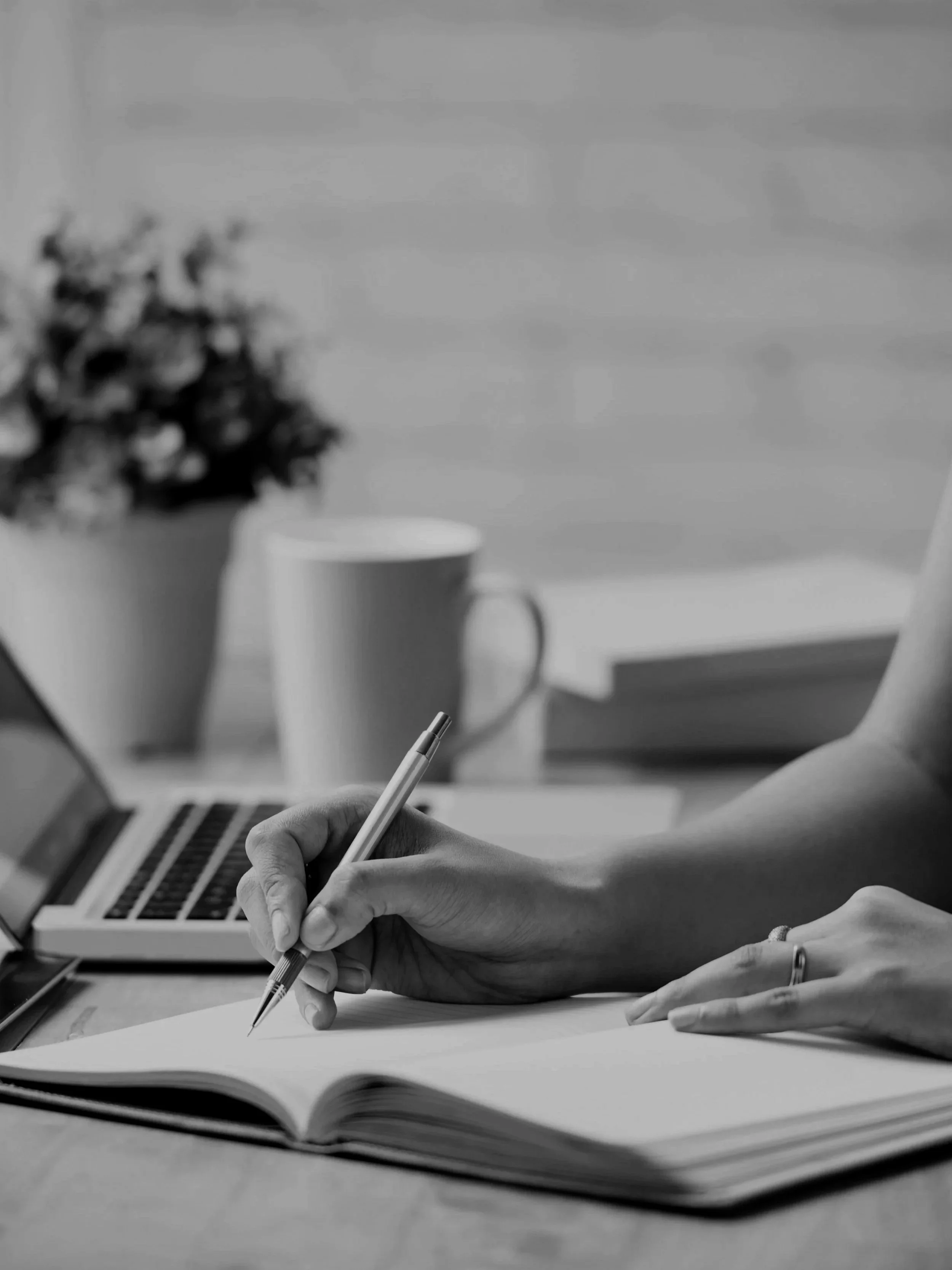 A person writes in a notebook with a pen, on a desk with a laptop, mug, plant, and books in the background.