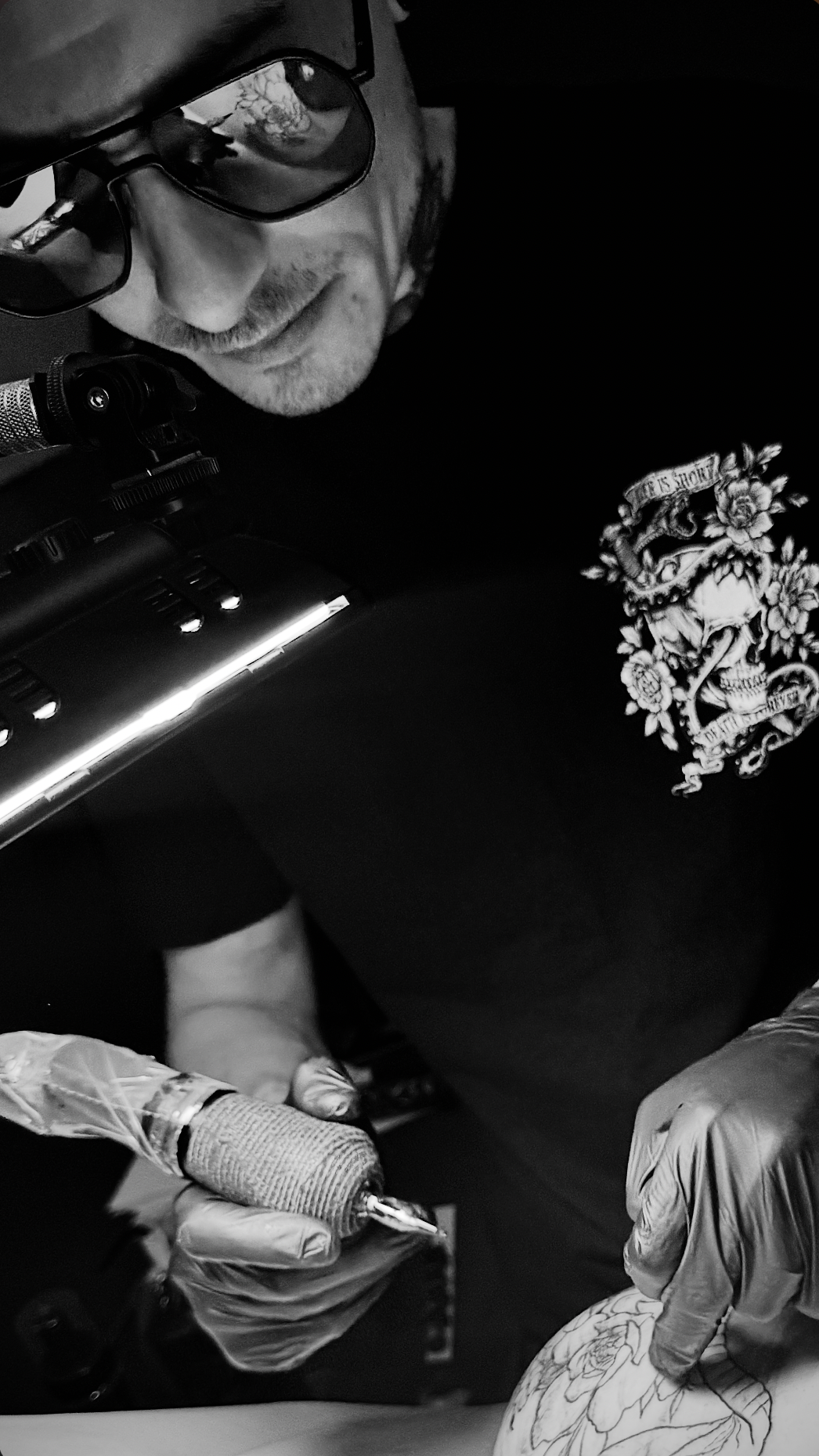 A man wearing glasses tattooing a floral design on a client’s arm in a tattoo studio, with tattoo equipment visible.